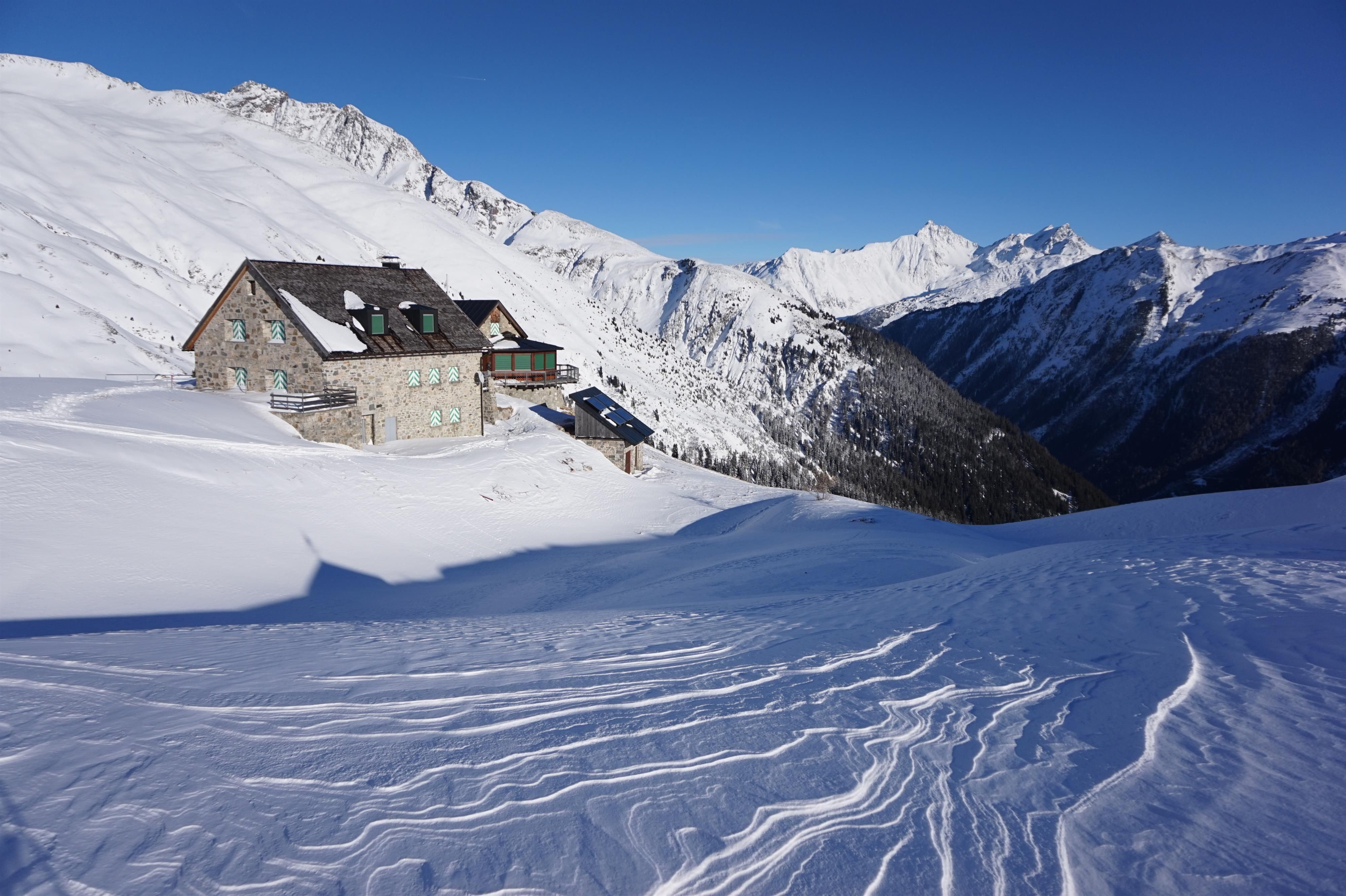 A picturesque mountain house in a snowy landscape. The surroundings are surrounded by tall mountains and clear skies.