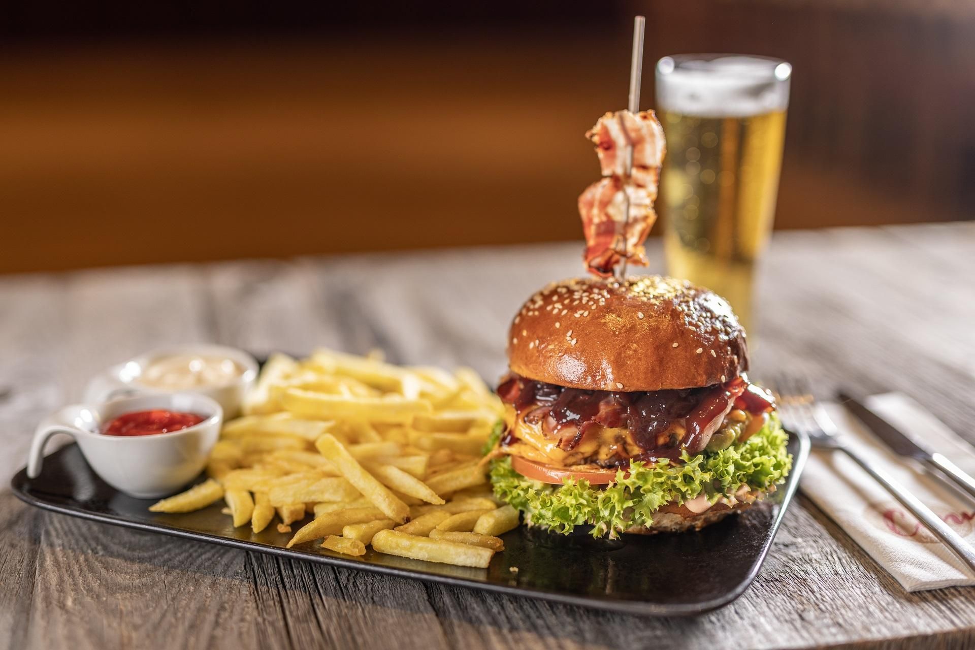 A delicious burger with bacon, lettuce, and a generous portion of French fries. Accompanied by a glass of beer on a rustic wooden table.
