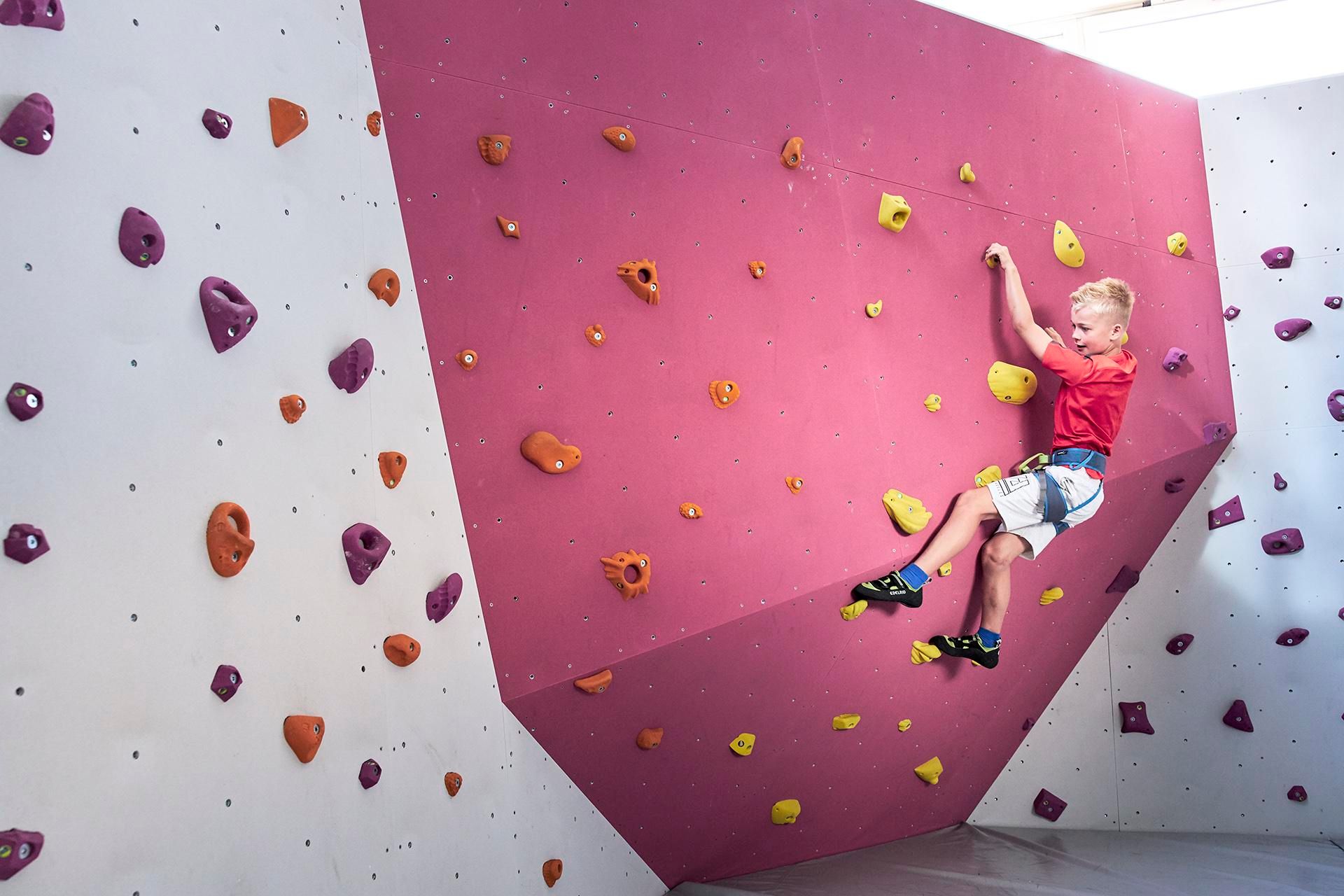 A boy is climbing on a pink and white climbing wall. The wall is equipped with colorful grips.