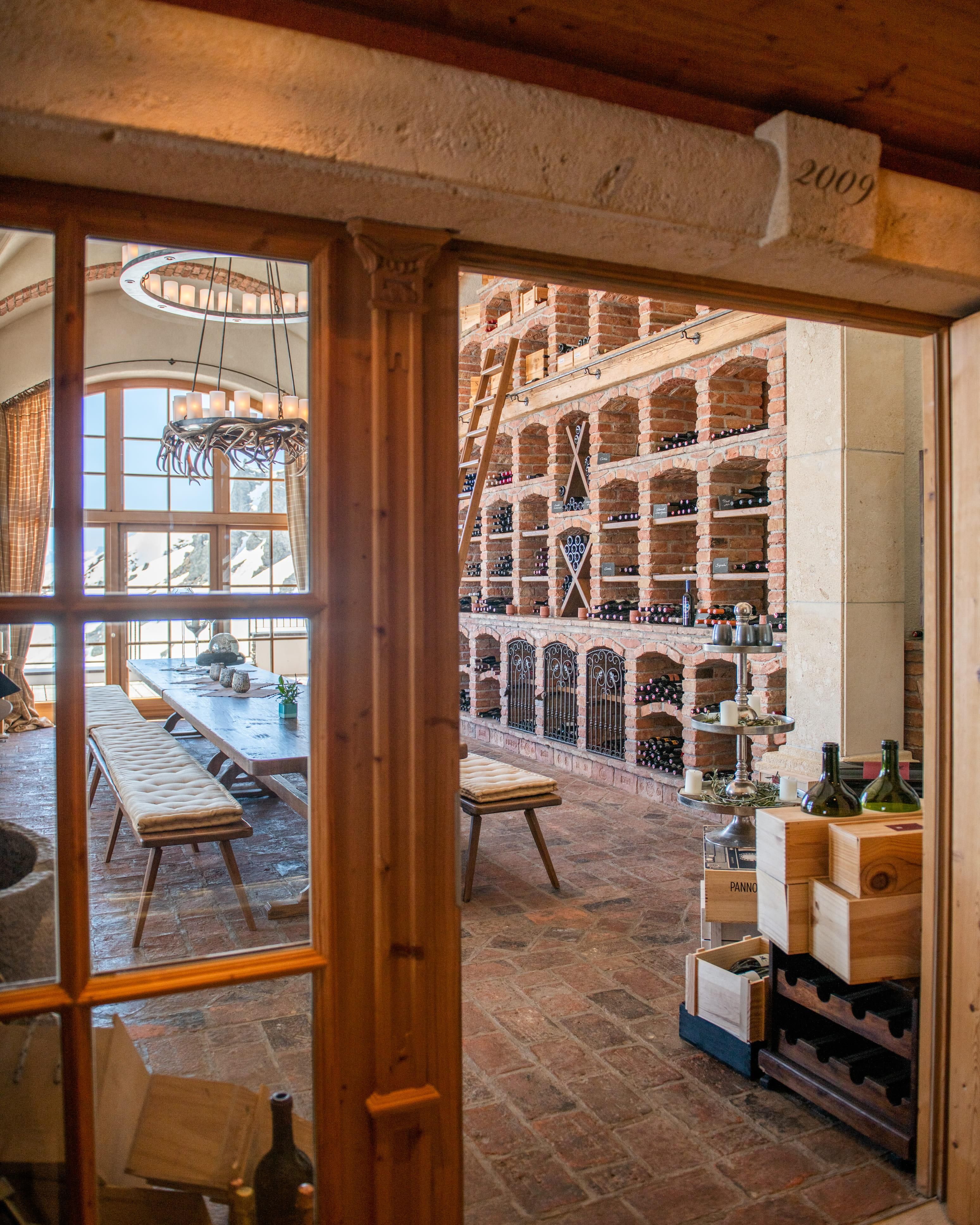 An elegant wine cellar with wooden shelves full of wine bottles. In the background, there is a long table with chairs and a large window front.