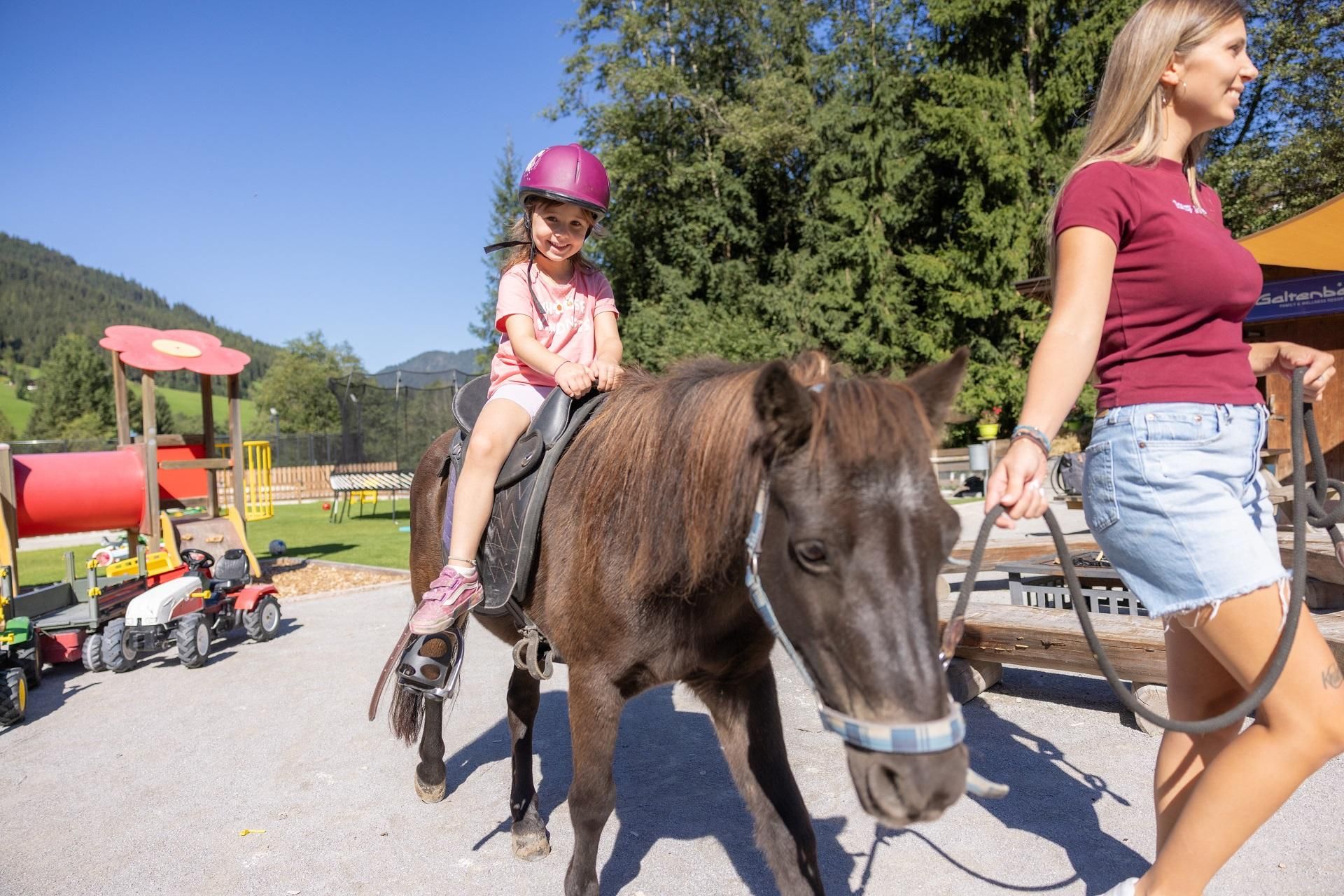 A little girl is riding on a pony and wearing a helmet. A woman is leading the pony in a playground with green surroundings.