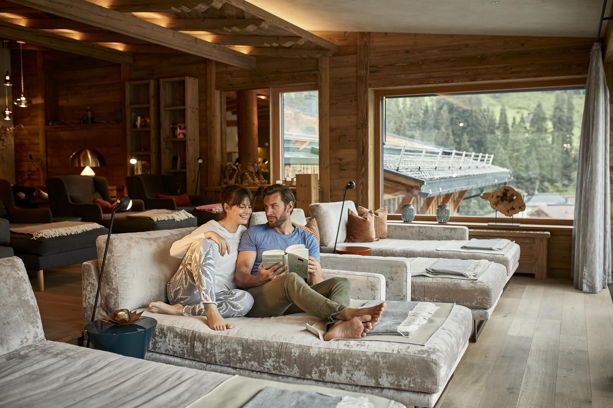 A cozy room with modern loungers and large windows that offer a view of the mountains. A couple is enjoying a relaxing moment together and reading a book.