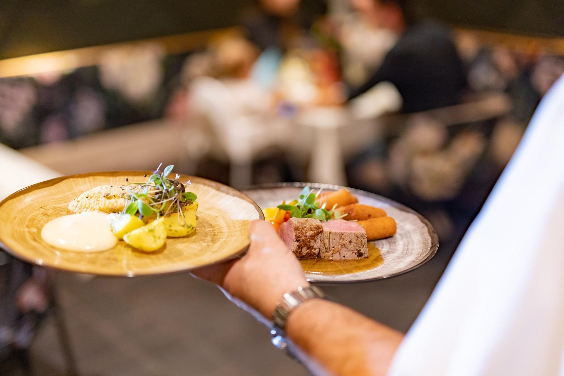 A waiter is carrying two plates of food. On the first plate are potatoes and vegetables, on the second plate are meat and side dishes.