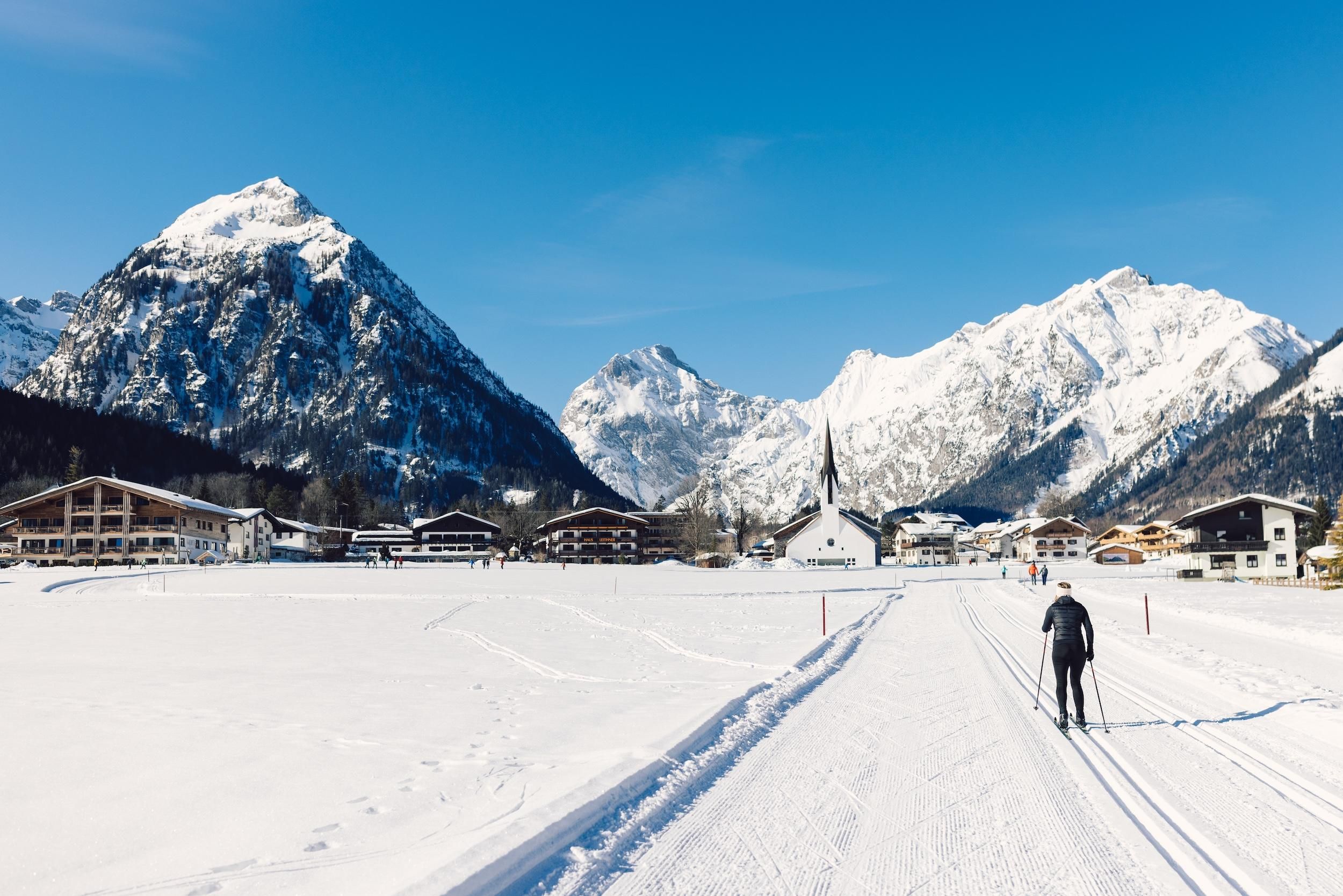A snowy landscape with tall mountains in the background. A person is cross-country skiing on the groomed trail.