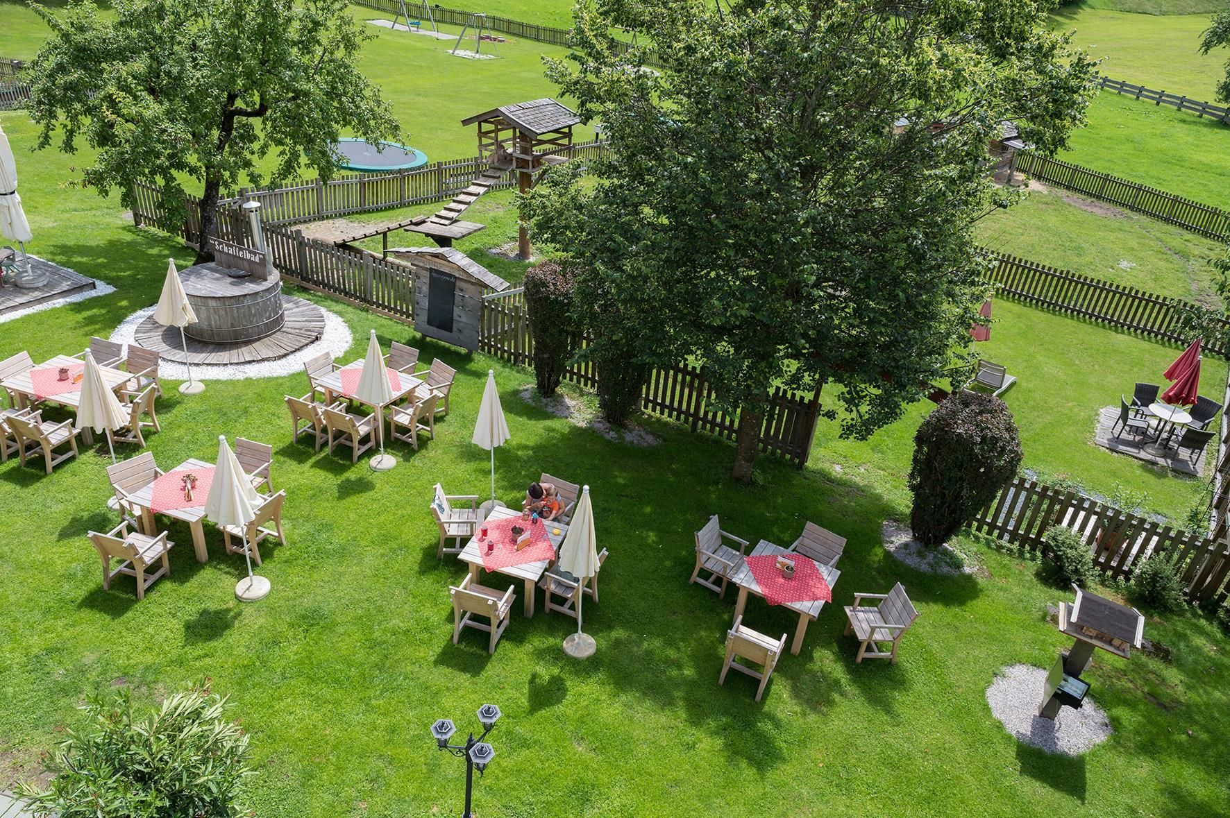 A cozy garden with tables and chairs under sun umbrellas. In the background, a green lawn and a playground are visible.