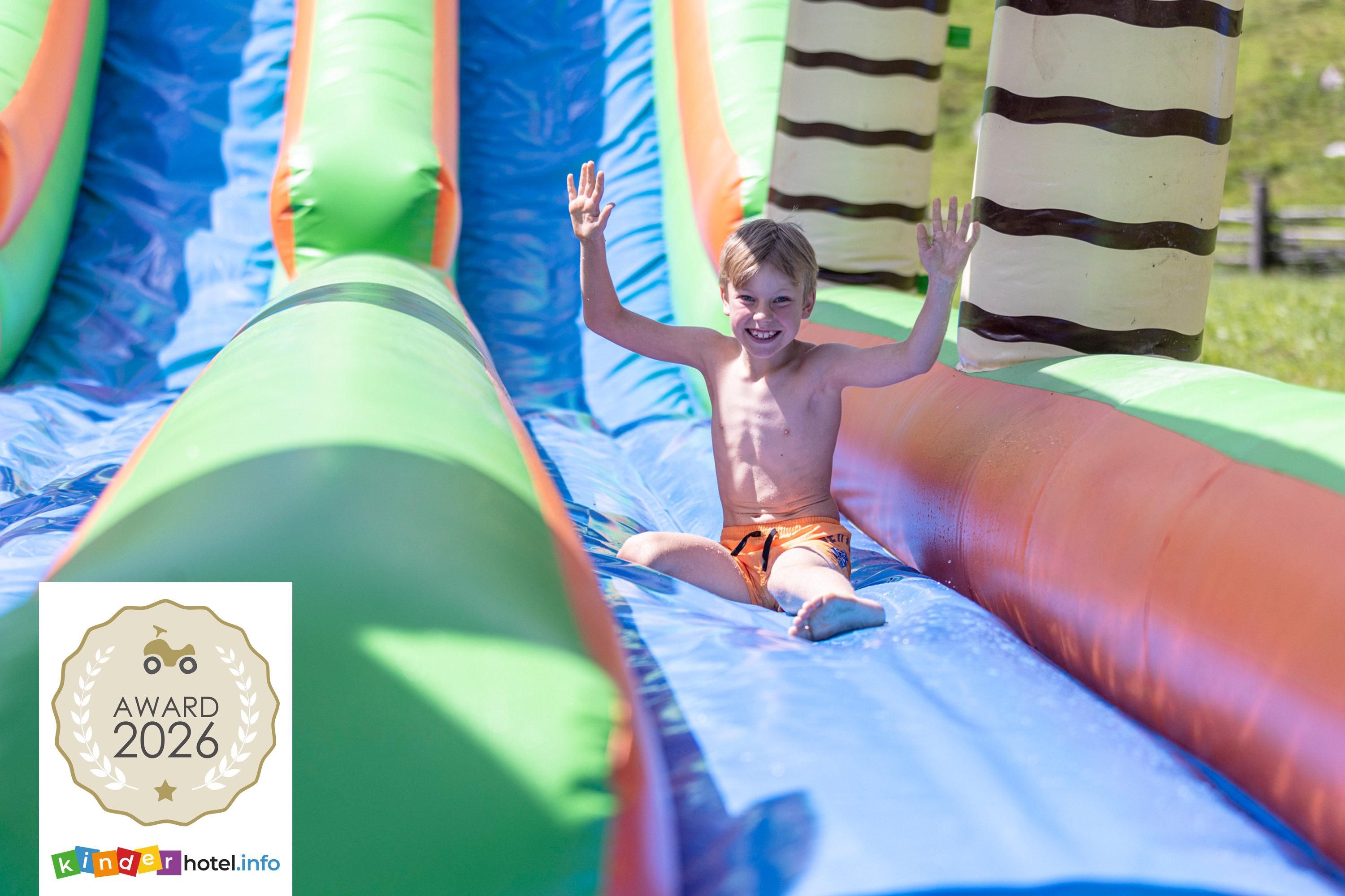 A boy happily slides down the water slide, lifting his hands in the air. In the background, there are colorful slide elements and a green meadow.