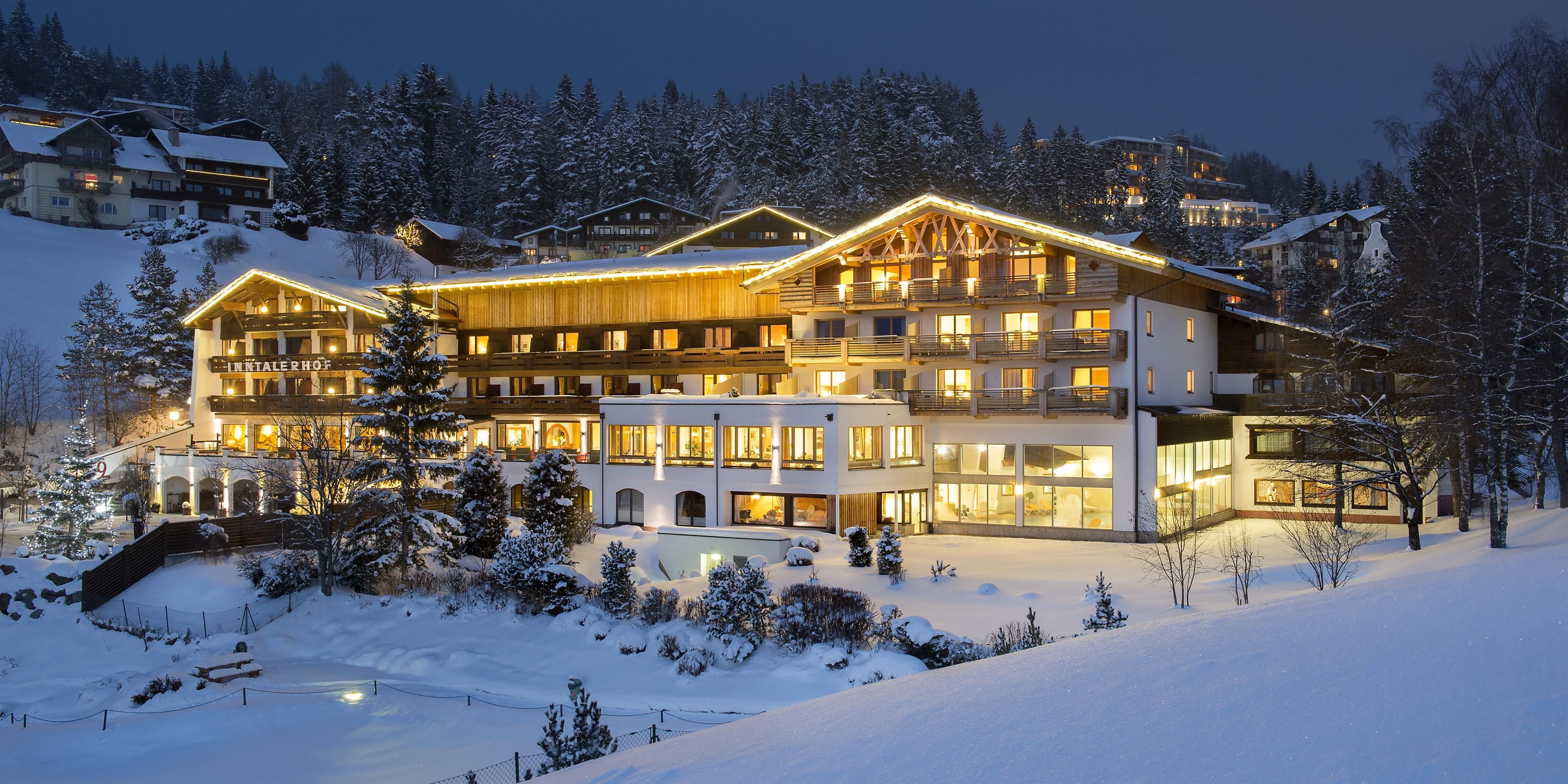 A winter hotel building illuminated in the evening. Surrounded by snow-covered trees and mountains, it radiates a cozy atmosphere.
