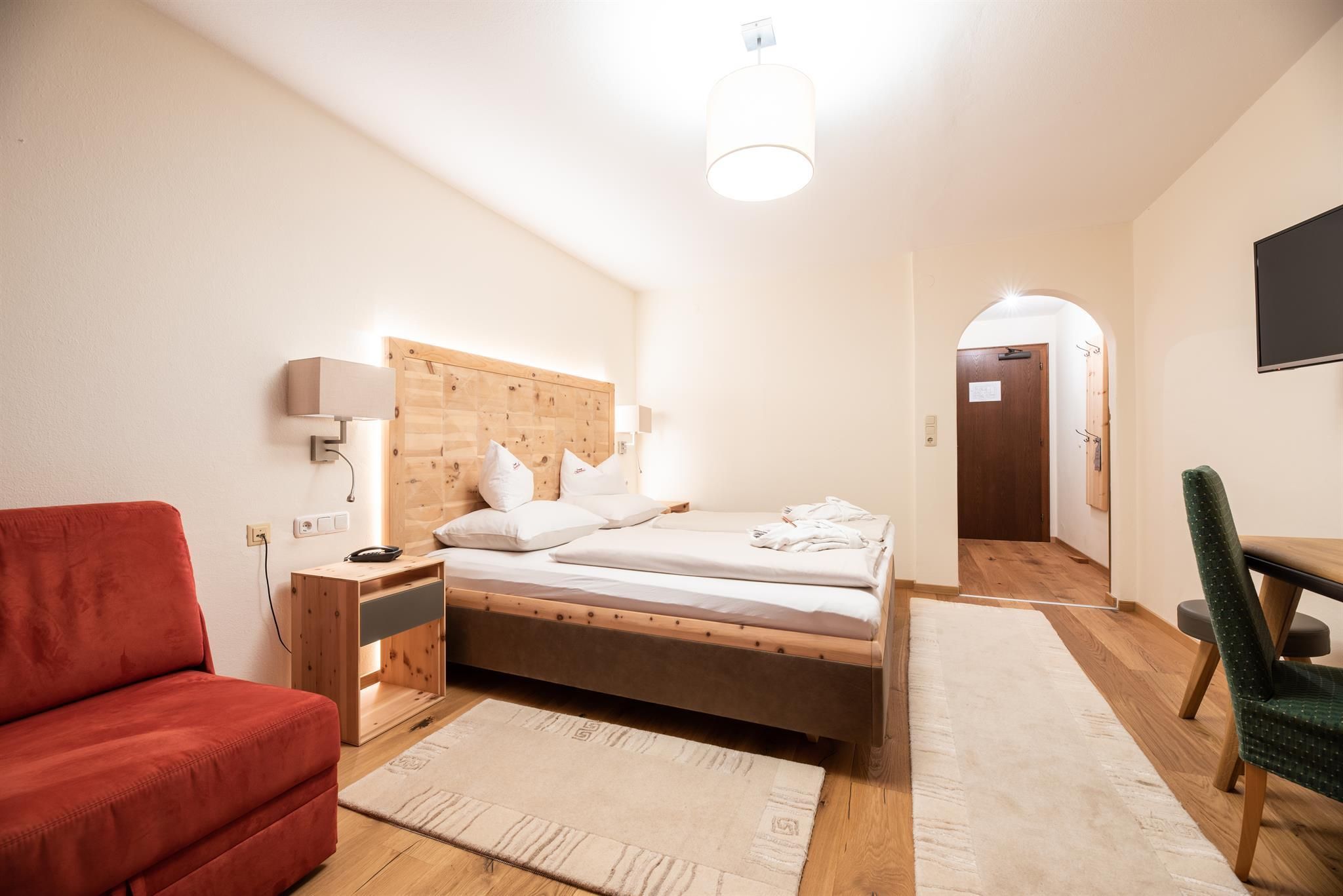 A cozy room with a double bed and a red armchair. The decor is modern and bright, with a wooden table and a television on the wall.
