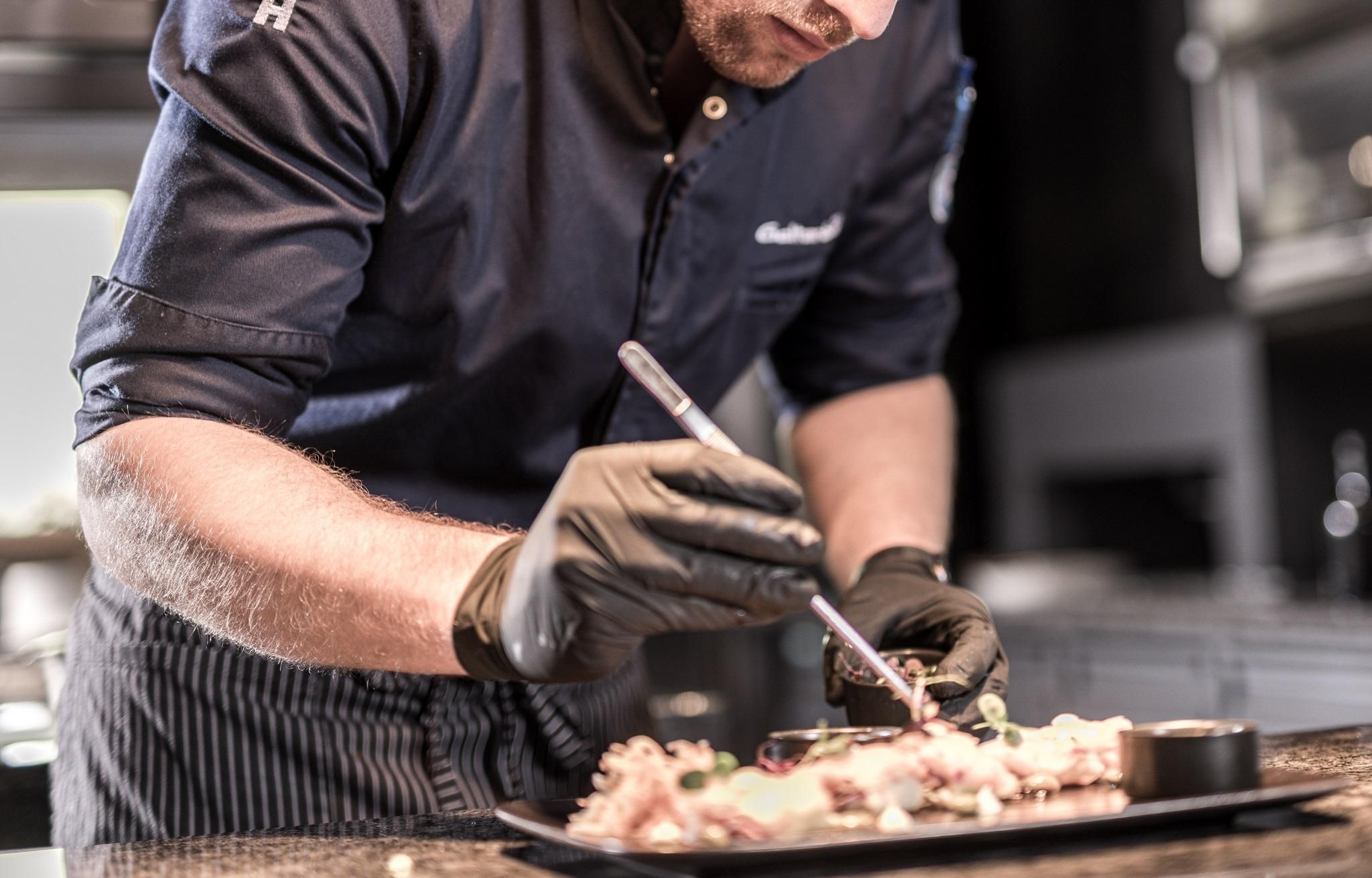 A chef is preparing a carefully arranged dish on a black plate. He is wearing gloves and focusing on the presentation of the food.