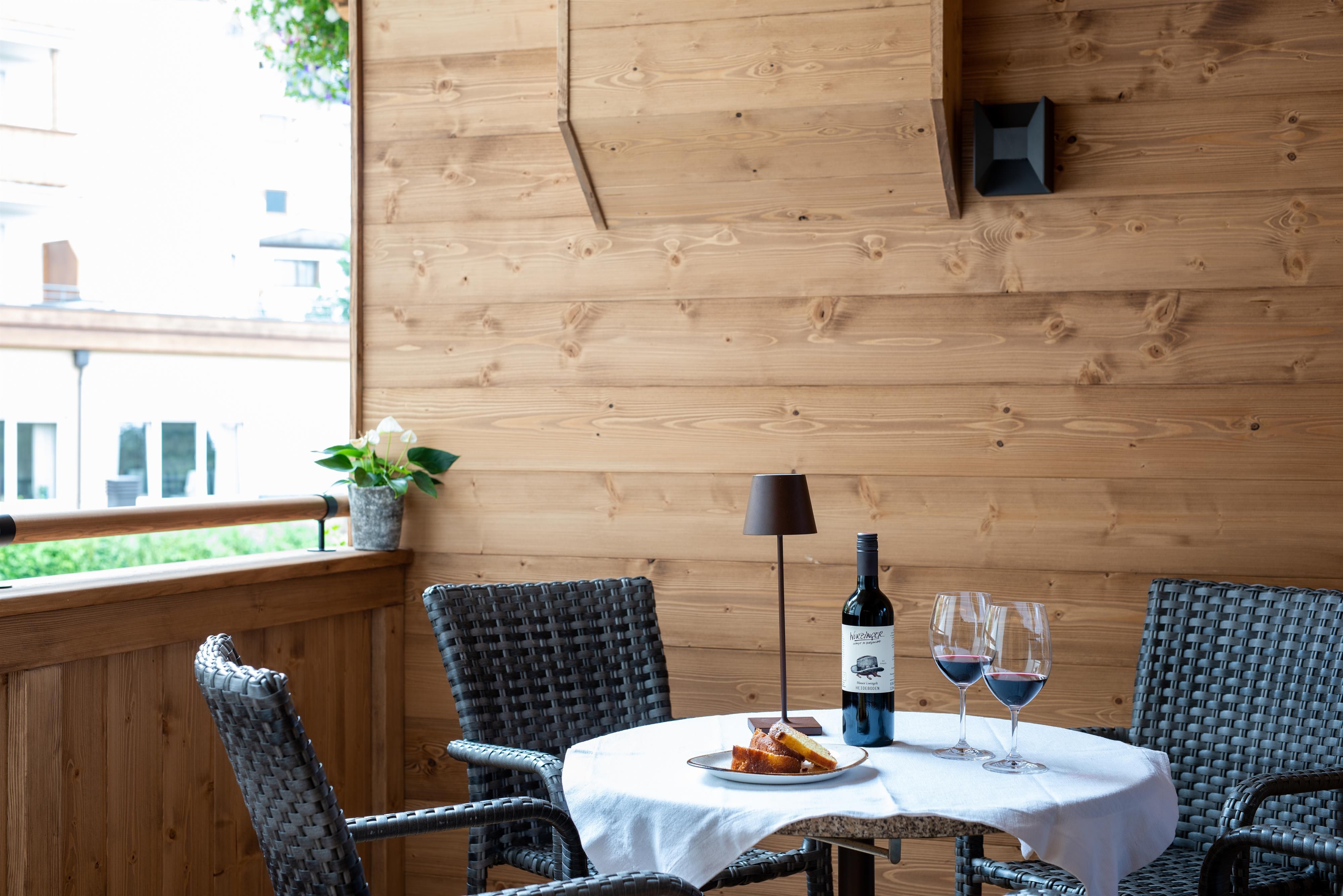 A cozy balcony with a small table, a bottle of wine, and glasses. The walls are made of wood, and there are plants in the background.