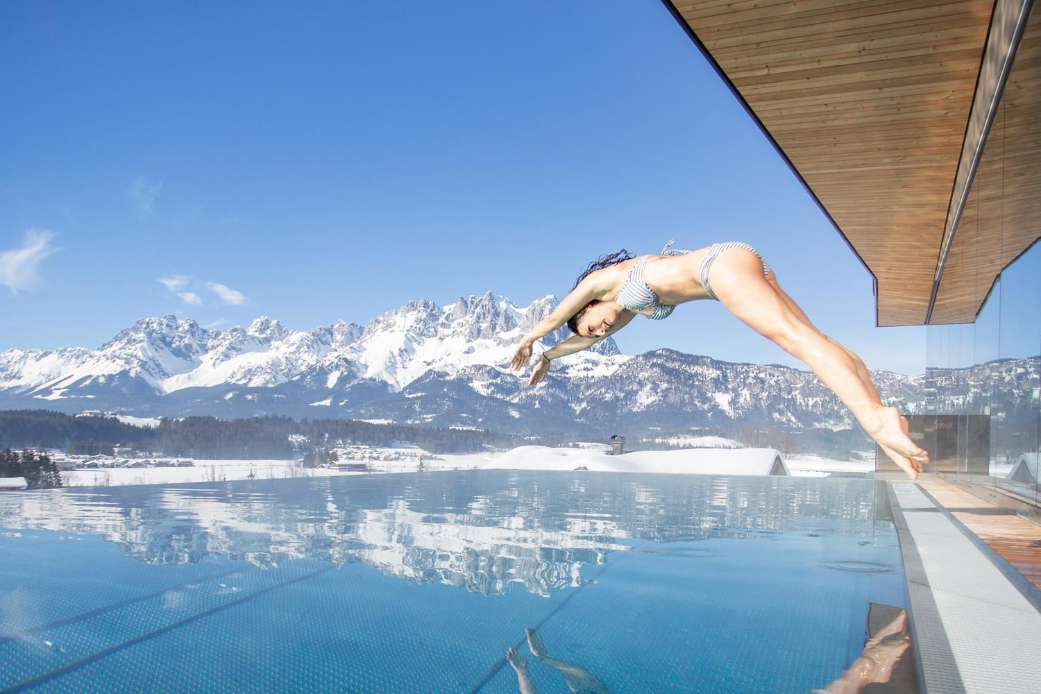 A person jumps off a platform into a pool with a view of the mountains. The sky is clear and blue, and the landscape is majestic.