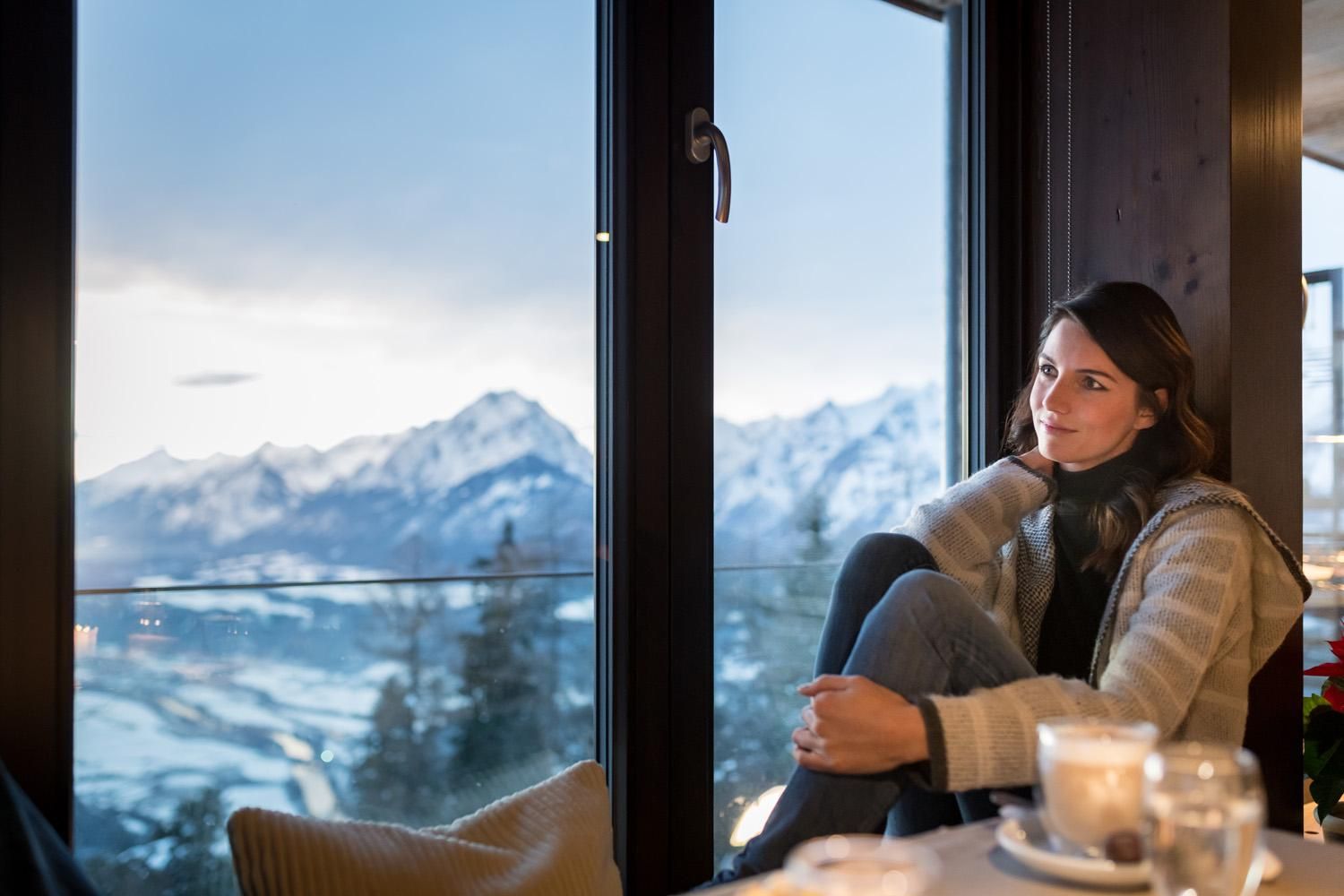 A woman sits by a window and gazes at the snow-covered mountains. The atmosphere is cozy and relaxing, with candlelight in the room.