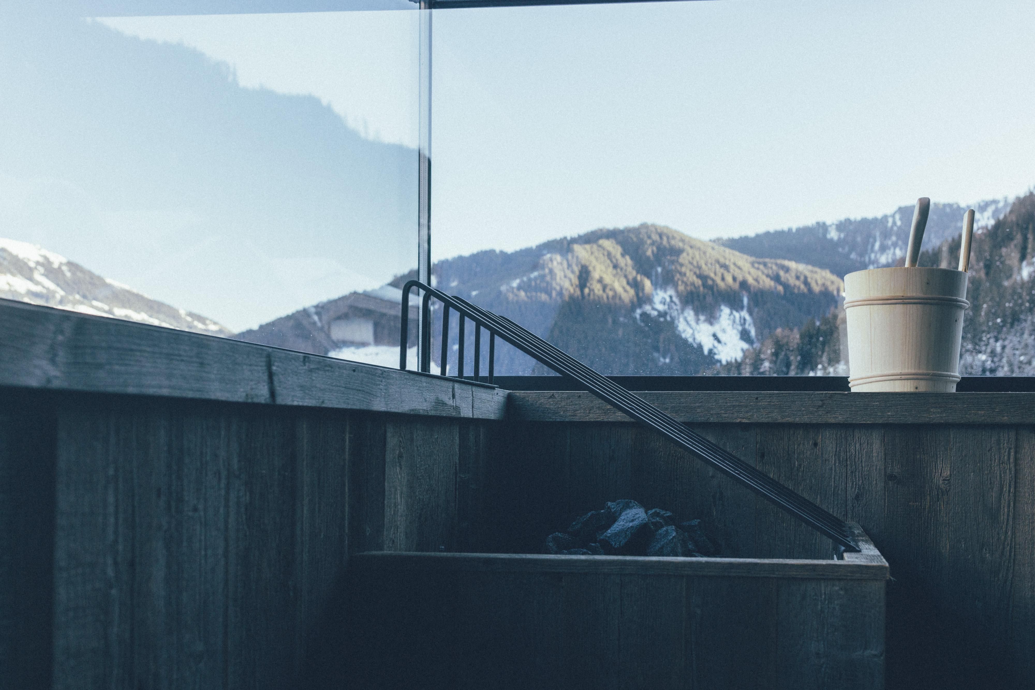 A cozy view from a wooden structure overlooking snow-covered mountains. Next to the window stands a container, probably for wellness.