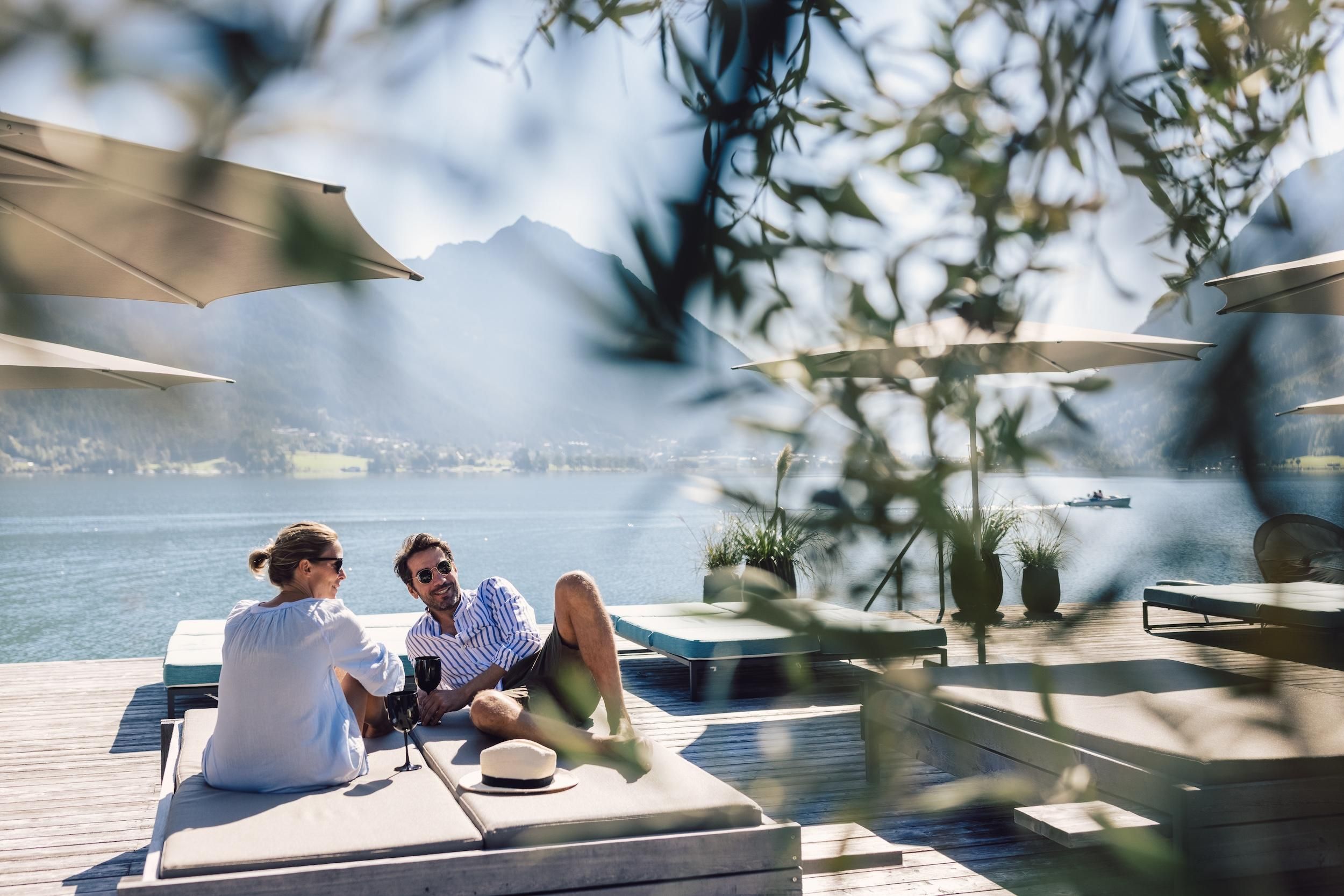 A relaxing moment by the water with two people on loungers. In the background, there are sun umbrellas and a beautiful view of the lake and the mountains.