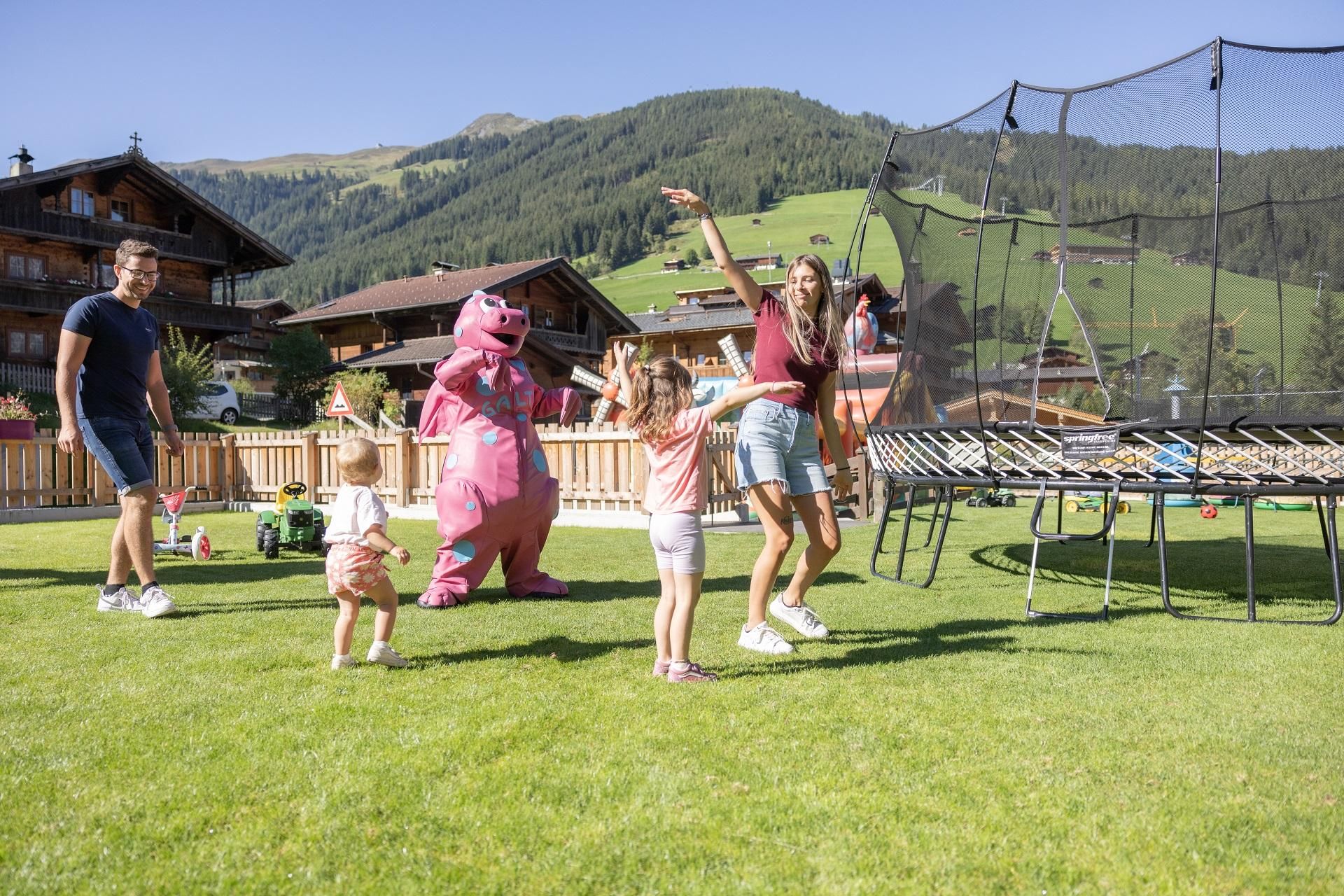 A cheerful group of people is dancing in the garden. In the background, a trampoline and picturesque mountains can be seen.