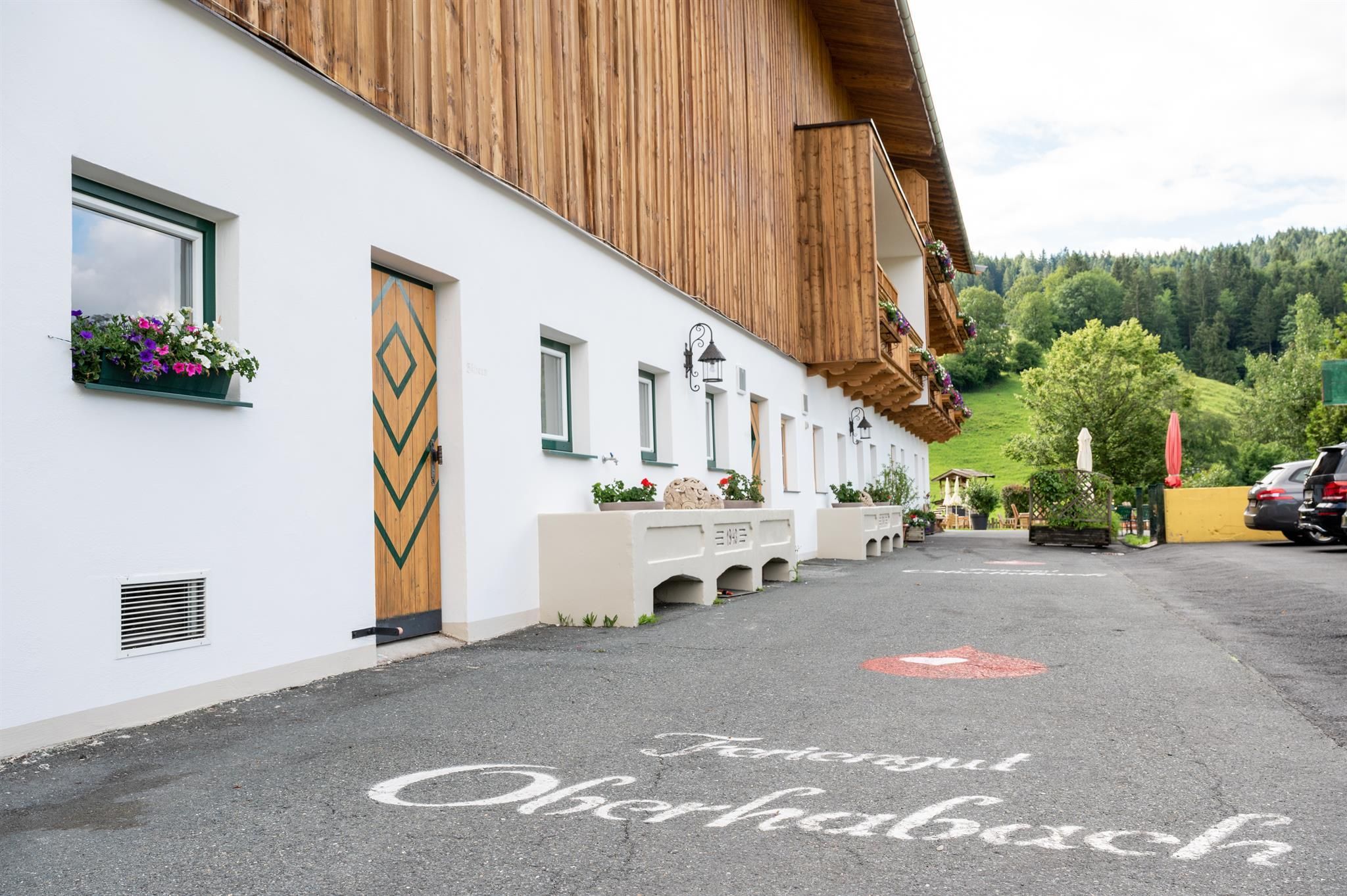 A cozy guesthouse with a wooden facade and blooming windows. The parking lot is tidy with a green environment in the background.