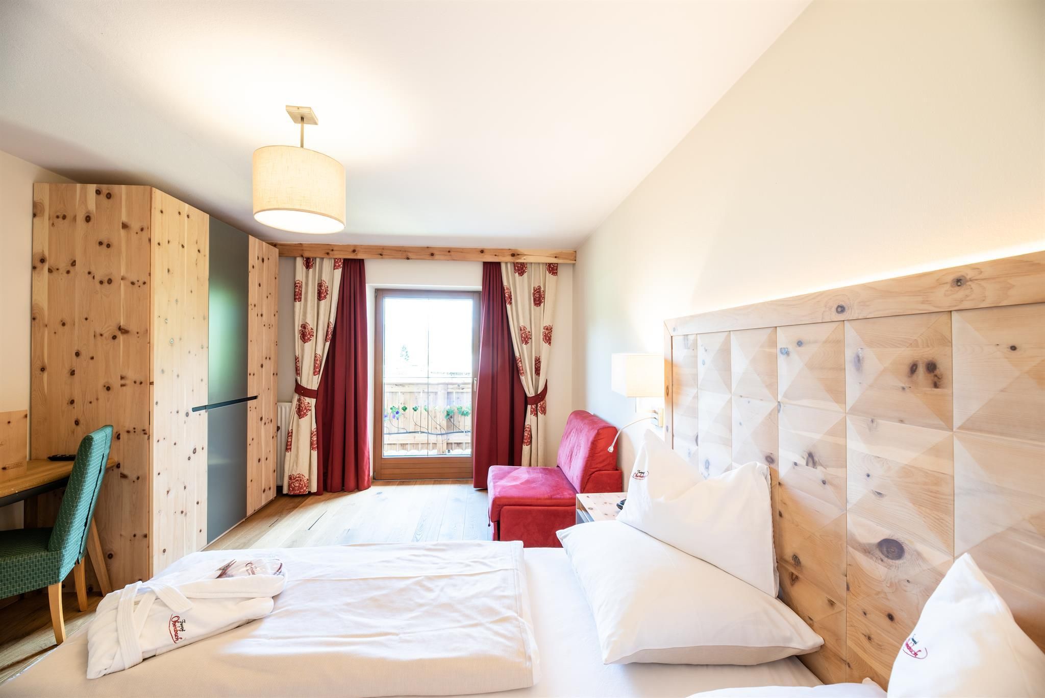 A cozy hotel room with wooden furniture and a large window. The room features a double bed, an armchair, and a small desk.