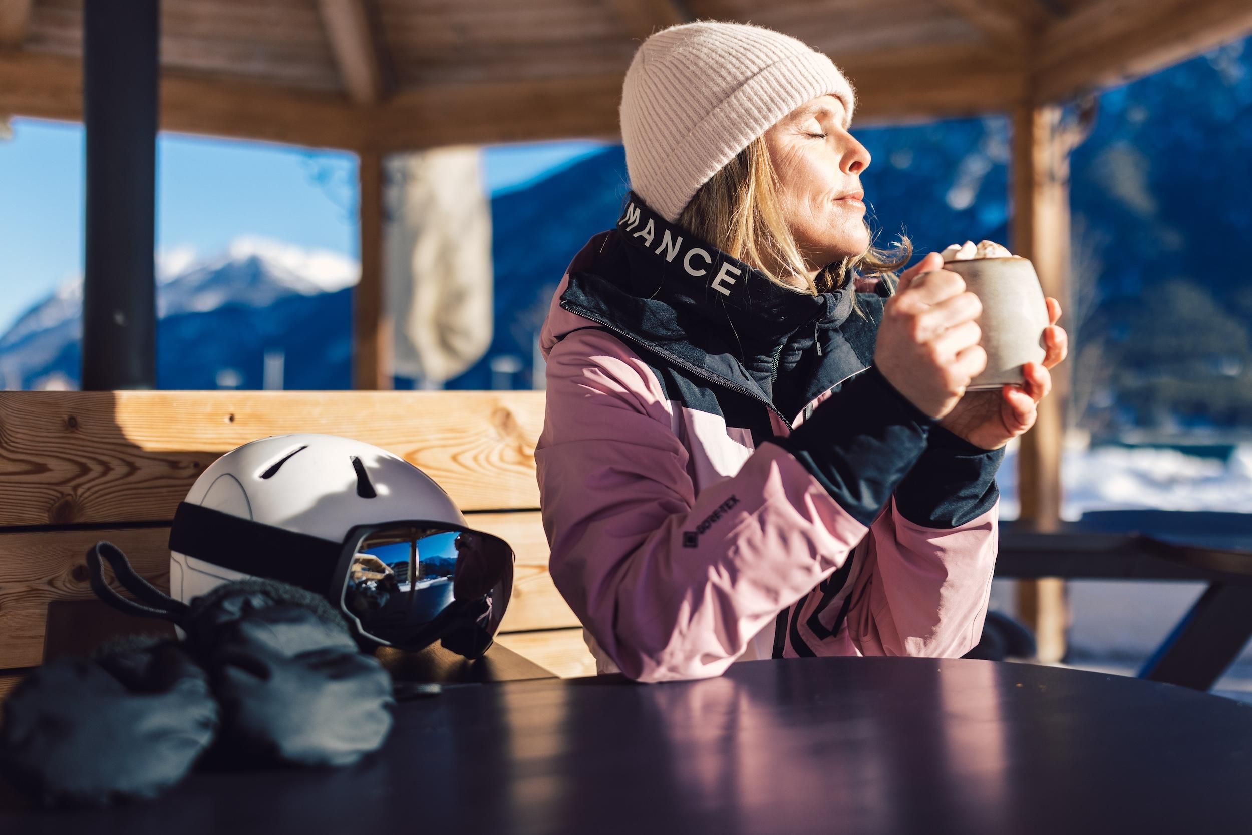 A woman is enjoying a warm drink in a mountain cabin. Next to her is a ski helmet and gloves on the table.