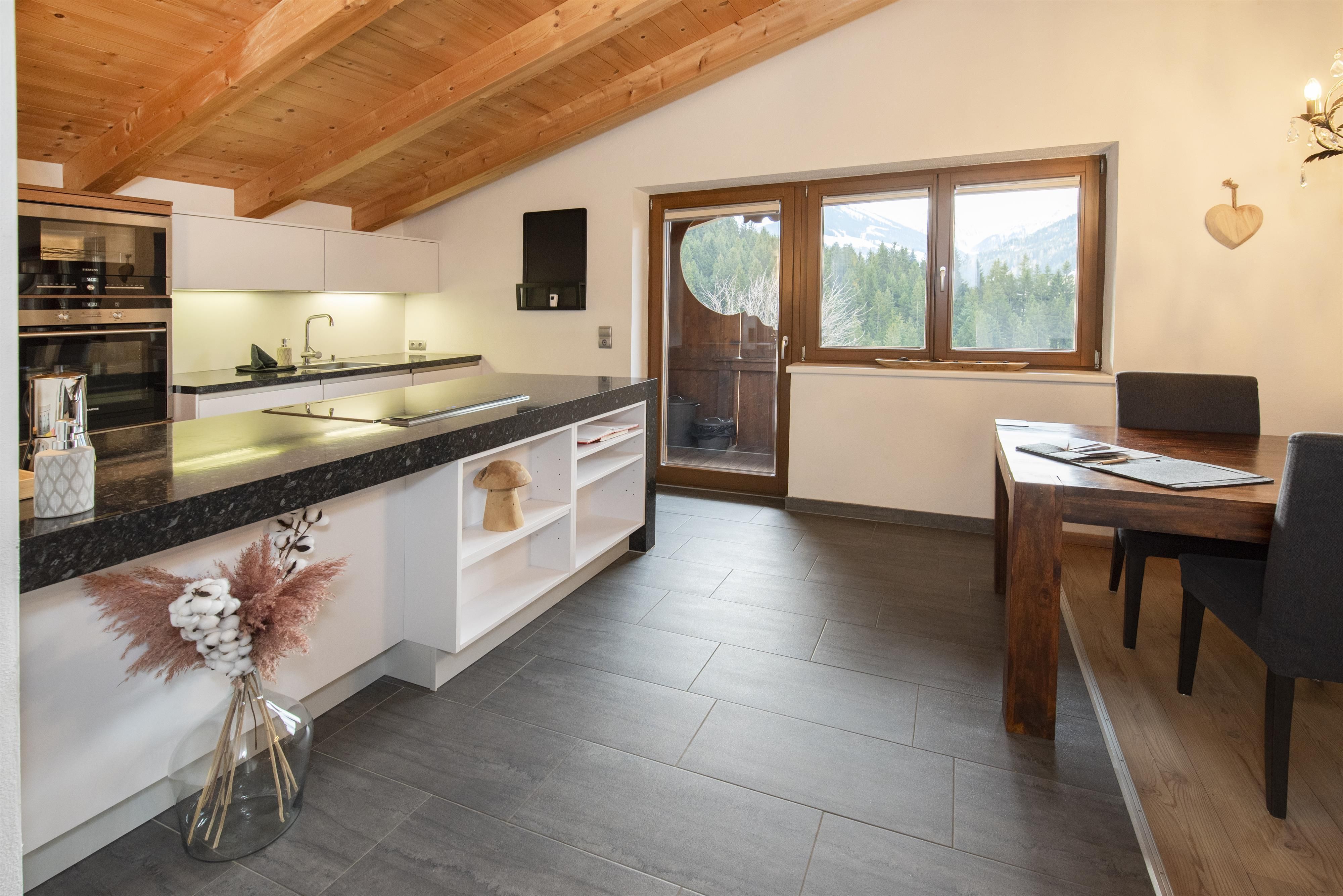 A modern kitchen with a wooden ceiling and elegant furniture. The room has large windows and offers a view of the outdoors.