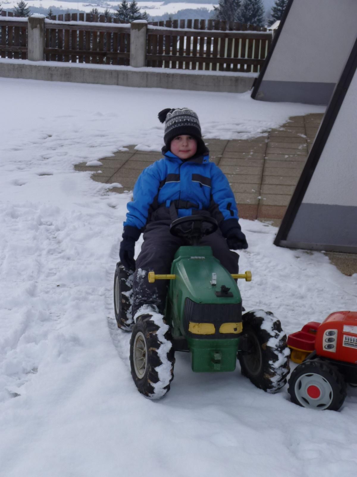 A boy is sitting on a green play tractor in the snow. He is wearing a blue anorak and a hat.