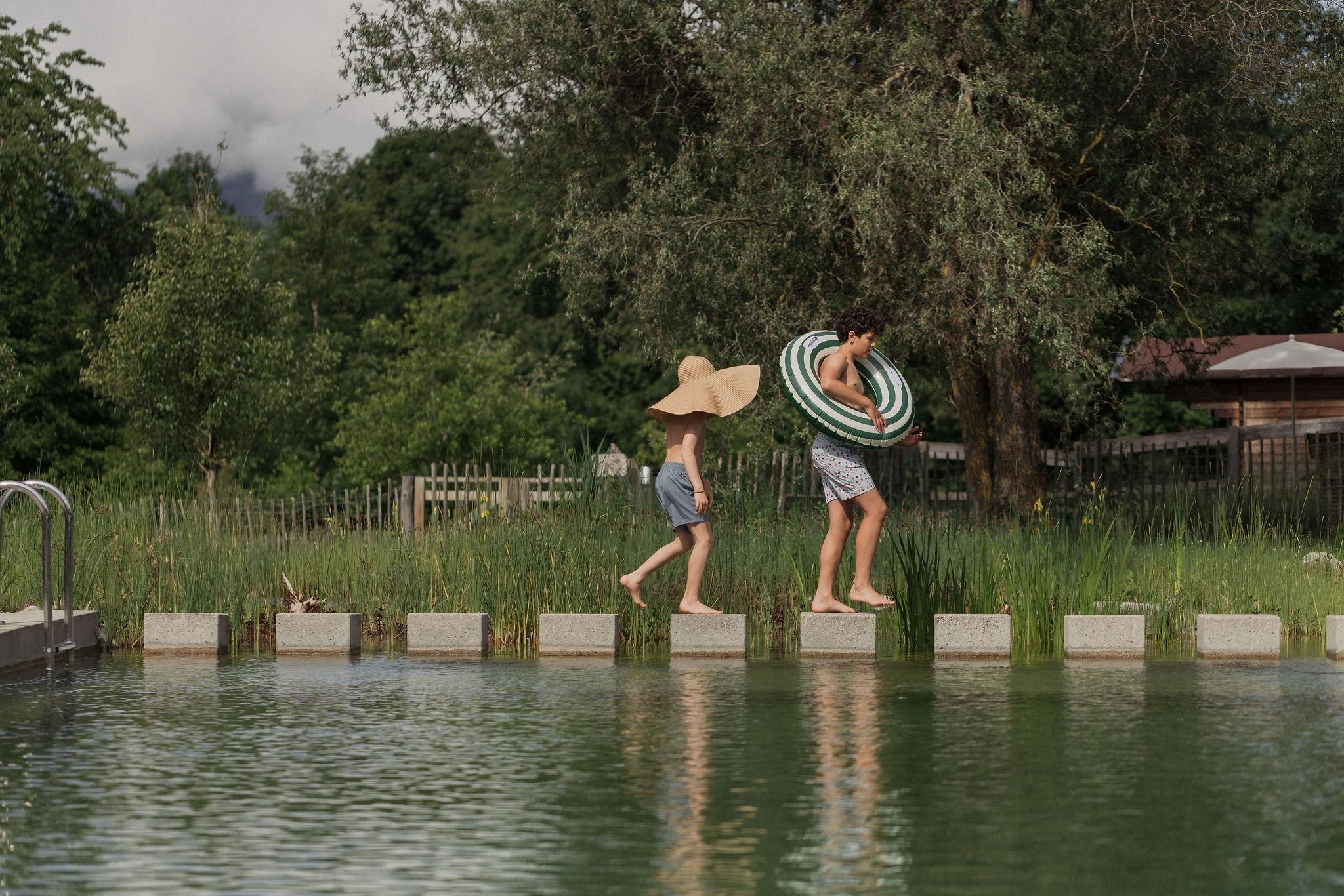 Two children are happily running along the edge of a swimming pool. One is wearing a large sun hat and the other a swimming ring.