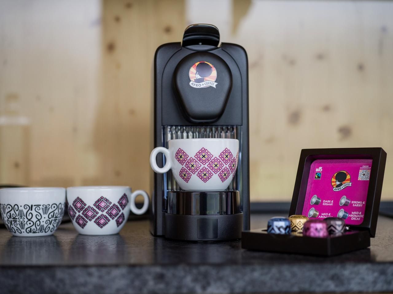 A coffee machine with a cup of coffee and colorful coffee capsules is on a table. Next to the machine are two decorative cups.