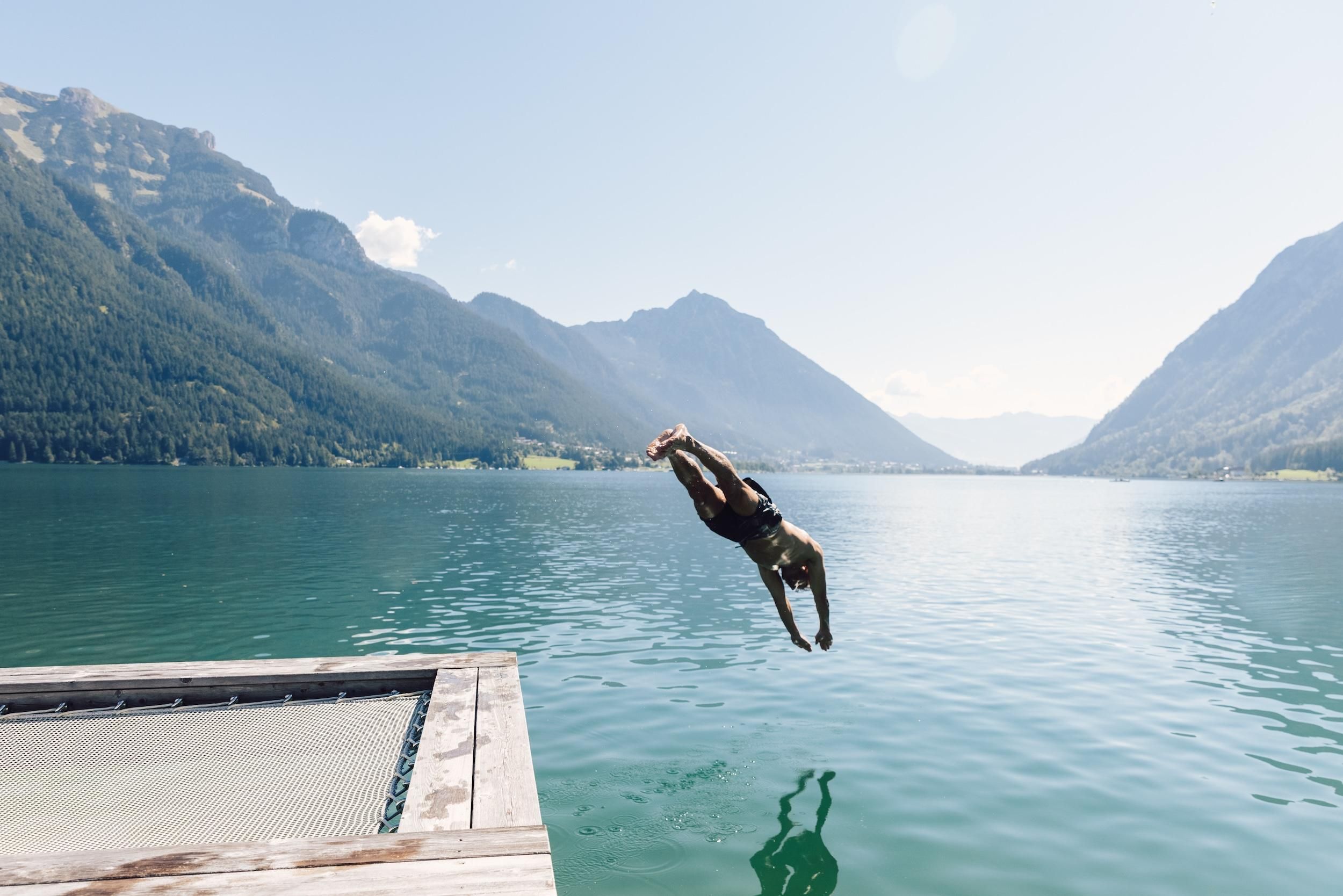 A man jumps from a dock into a clear, blue lake. In the background, majestic mountains and a bright blue sky can be seen.