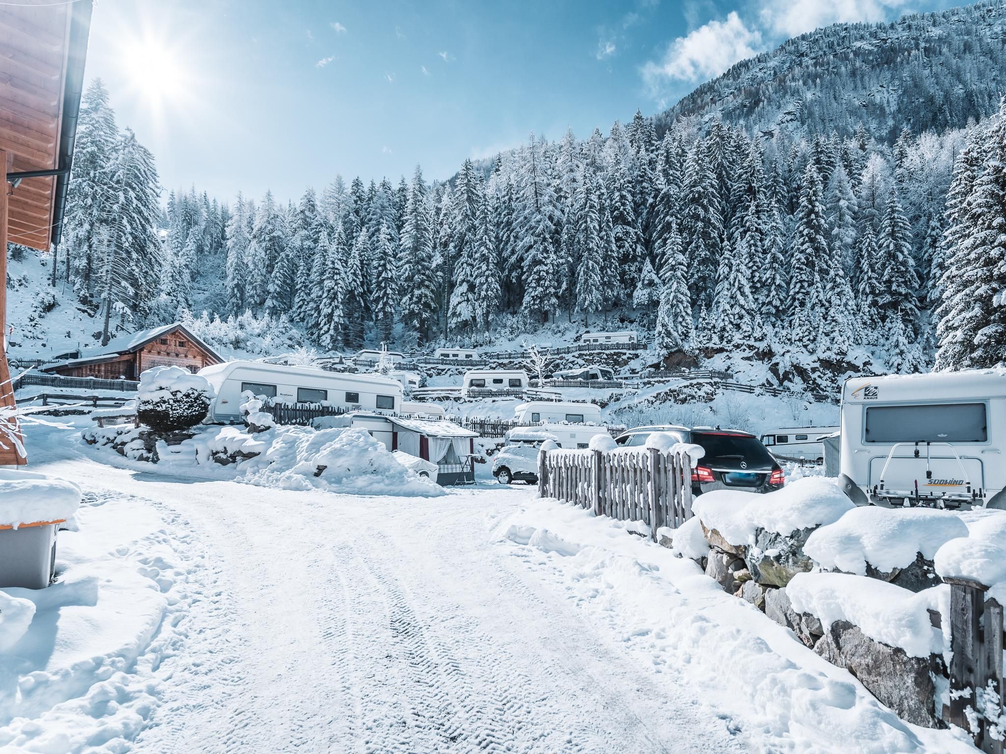 A snowy landscape with tall fir trees and a sun shining in the sky. In the foreground, a snow-covered path and some vehicles are visible.