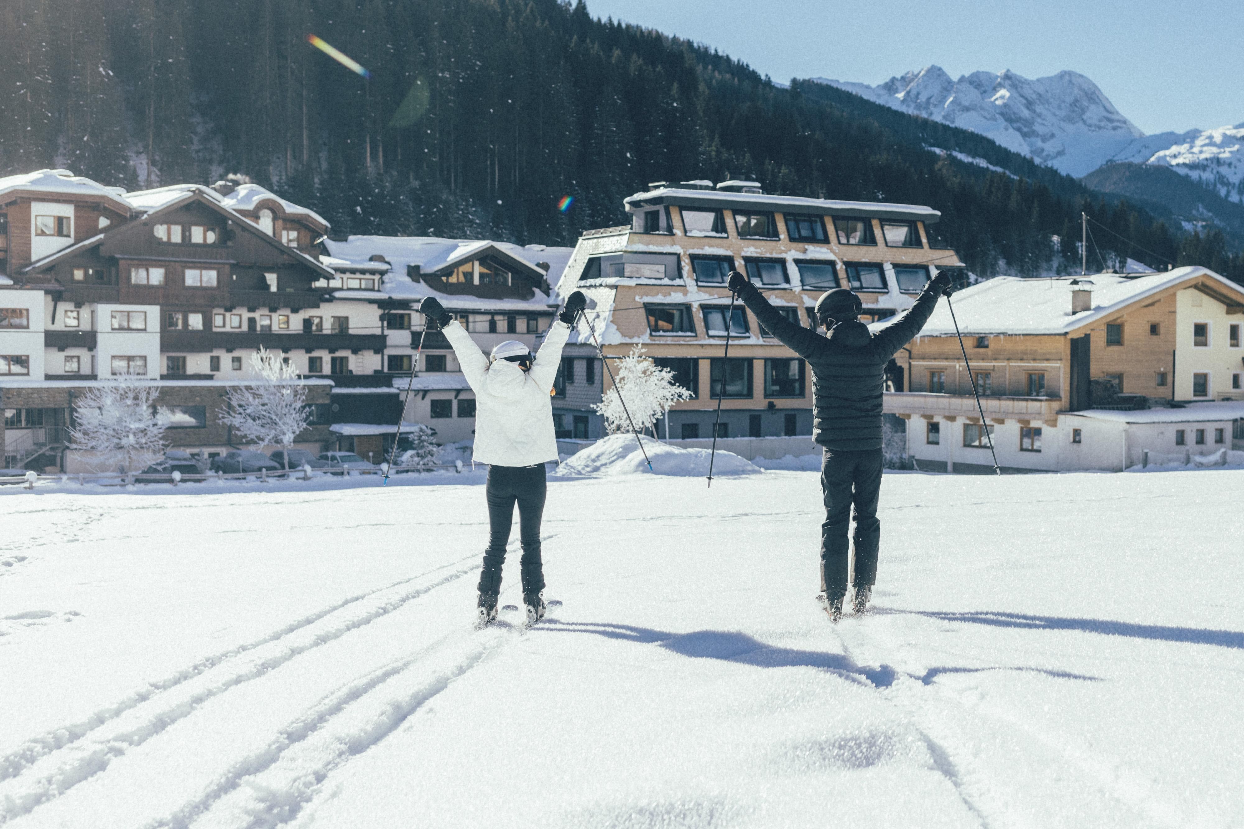 Two people are standing in the snowy landscape and are happy. In the background, alpine buildings and mountains can be seen.