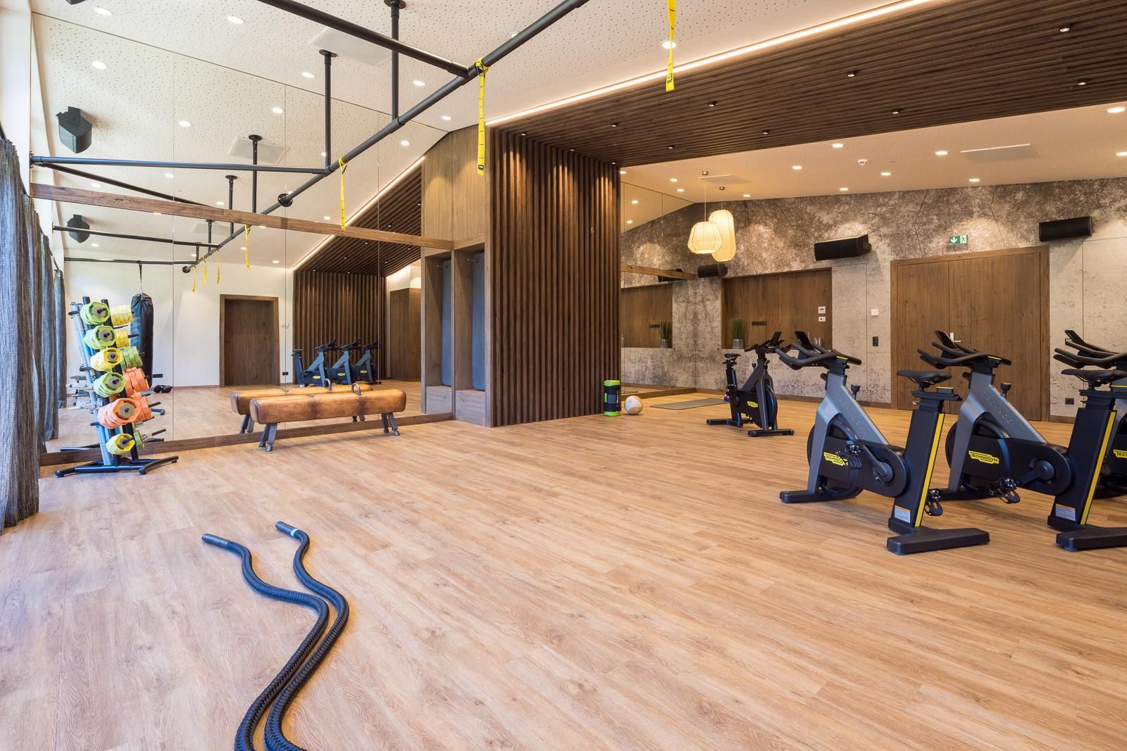 A modern fitness room with wooden flooring and mirror walls. The equipment includes bikes and various fitness machines.