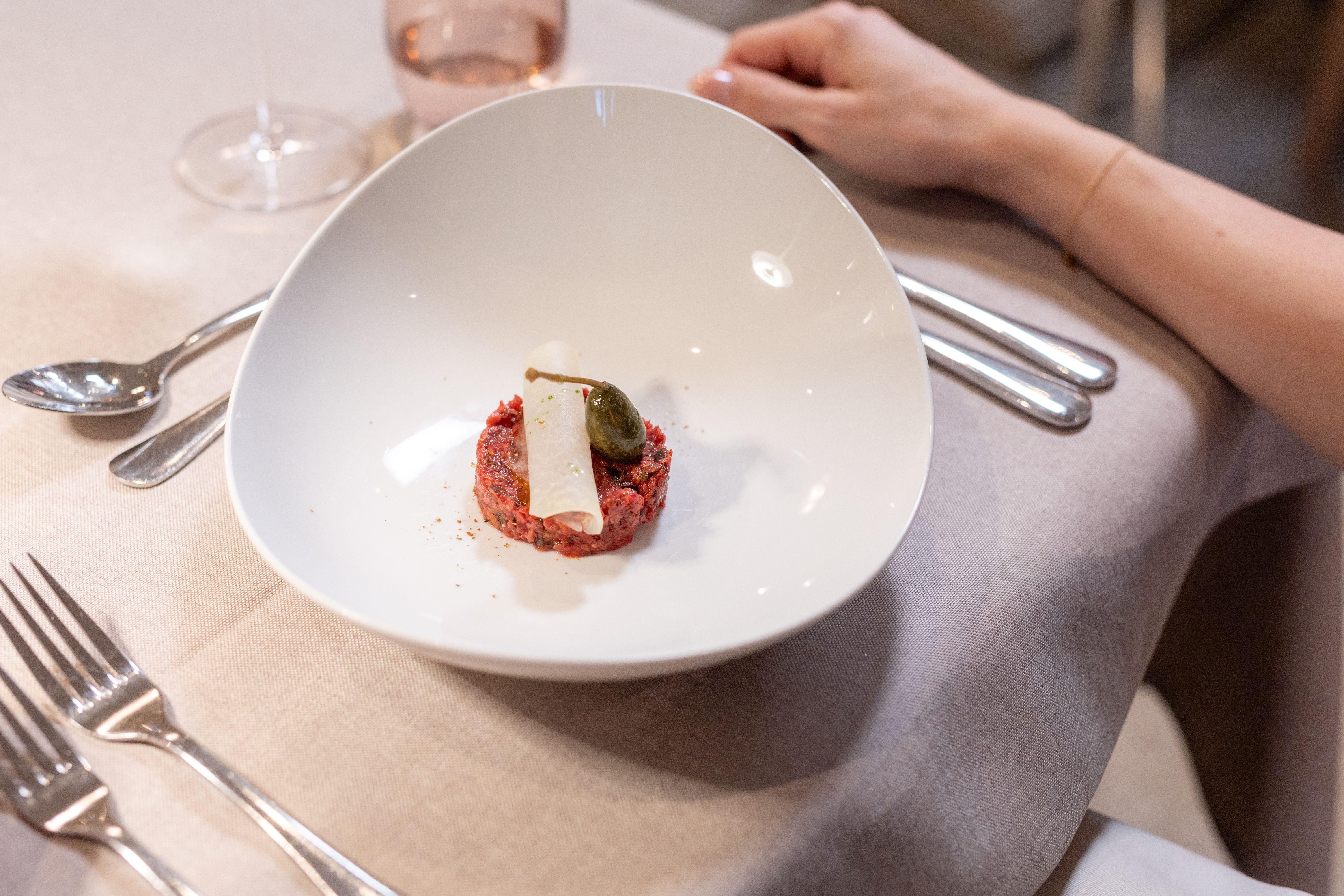 An elegant dish on a white plate, consisting of raw meat with a slice of cheese and an olive. In the background, cutlery and a glass are visible.