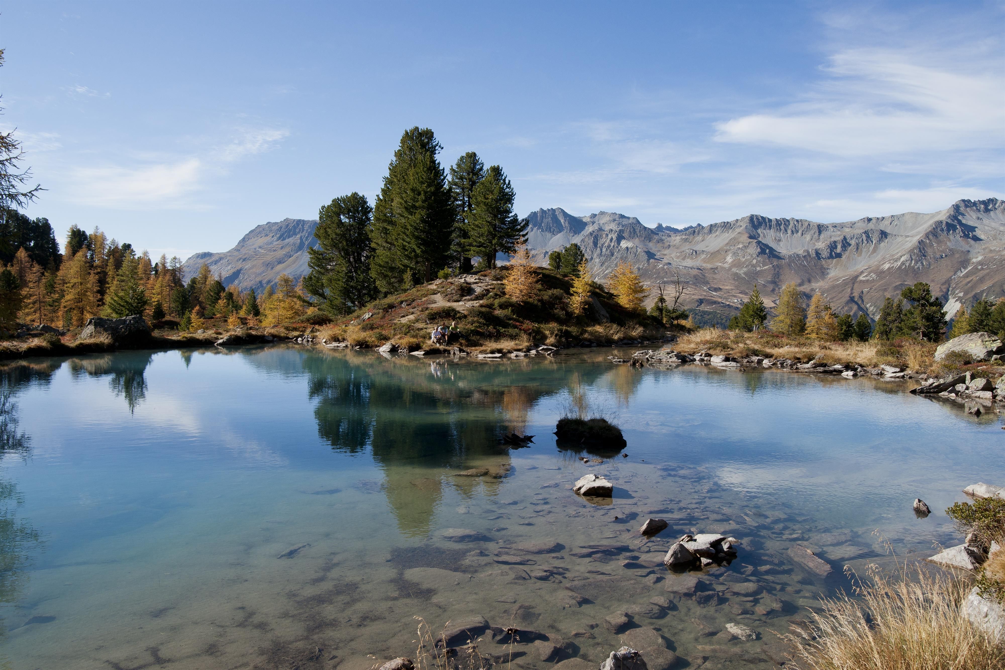 A clear mountain lake surrounded by green trees and majestic mountains. The calm water surface reflects the surroundings.