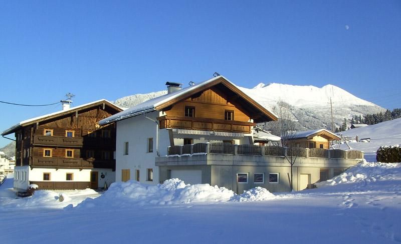 A picturesque house in a snowy landscape with mountains in the background. The sky is clear and blue.