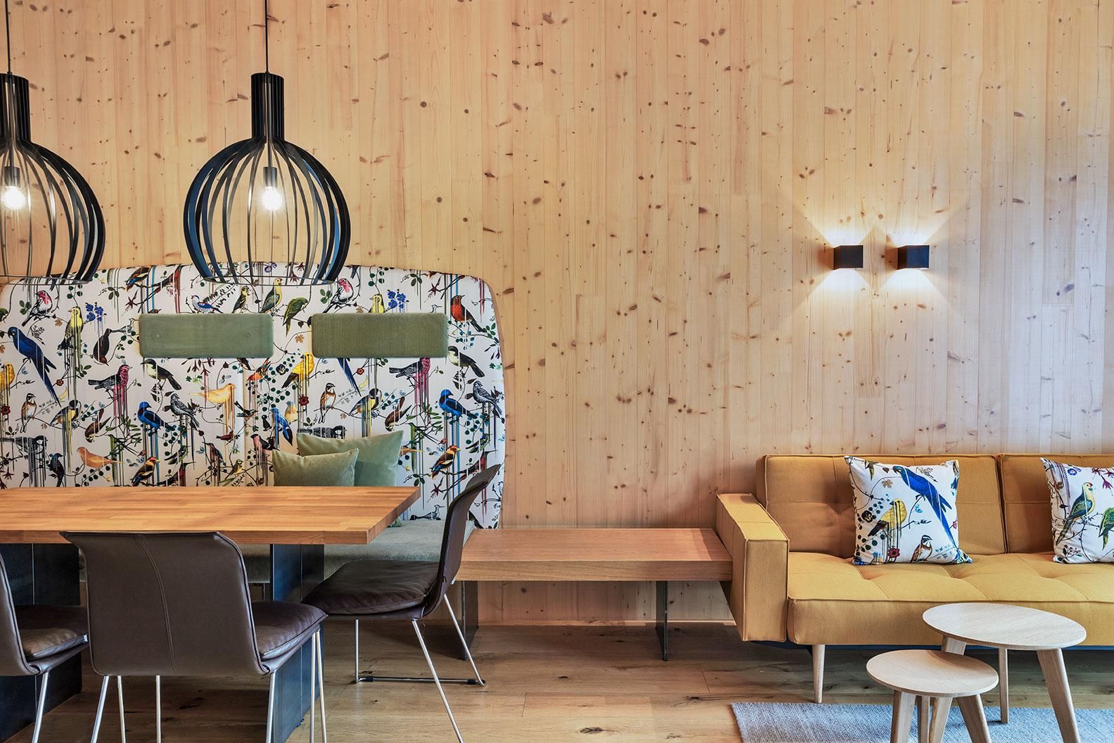 A modern living room with warm wood paneling. The room features a cozy yellow sofa, a dining area, and stylish lamps.