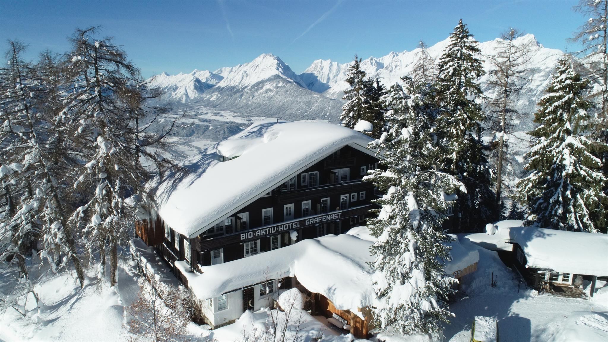 A picturesque chalet in the snow, surrounded by tall, snow-covered trees. In the background, majestic mountains stretch beneath a clear blue sky.