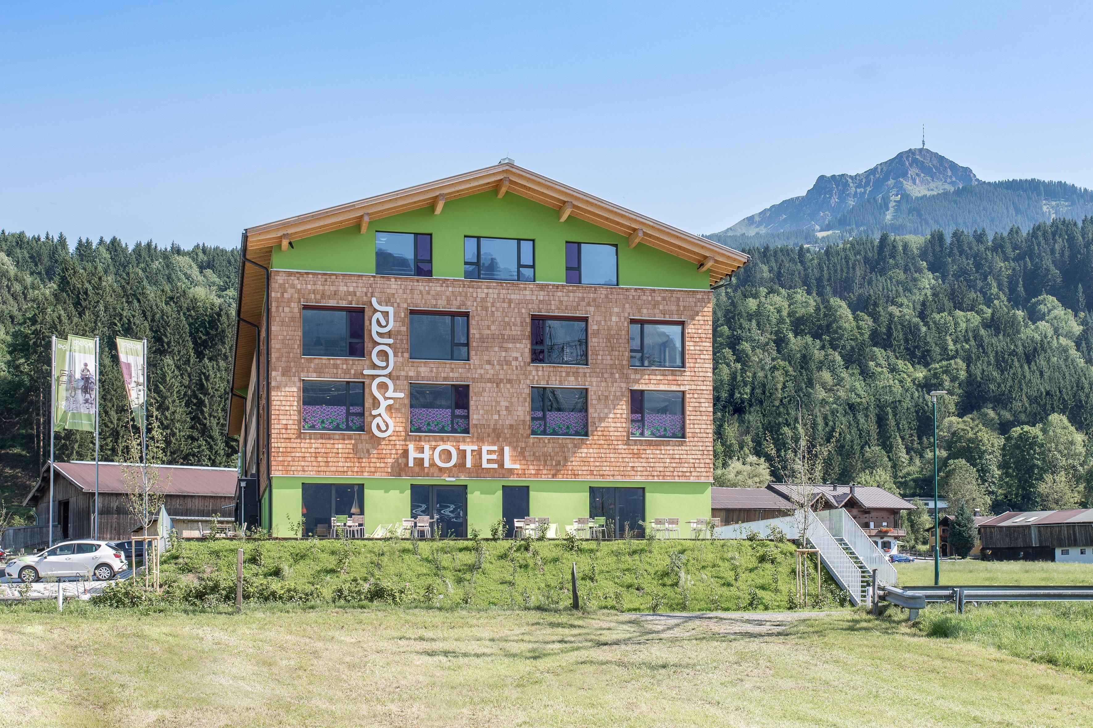 A modern hotel building with a colorful design and large windows. In the background, forests and mountains are visible, providing a beautiful natural backdrop.
