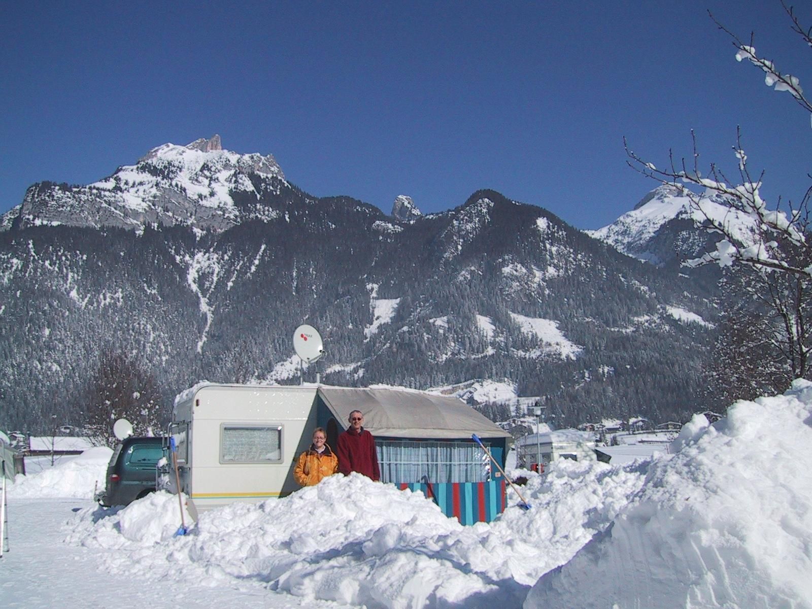 A winter landscape with snow-covered mountains in the background. In front of a caravan, two people are standing in the snow.
