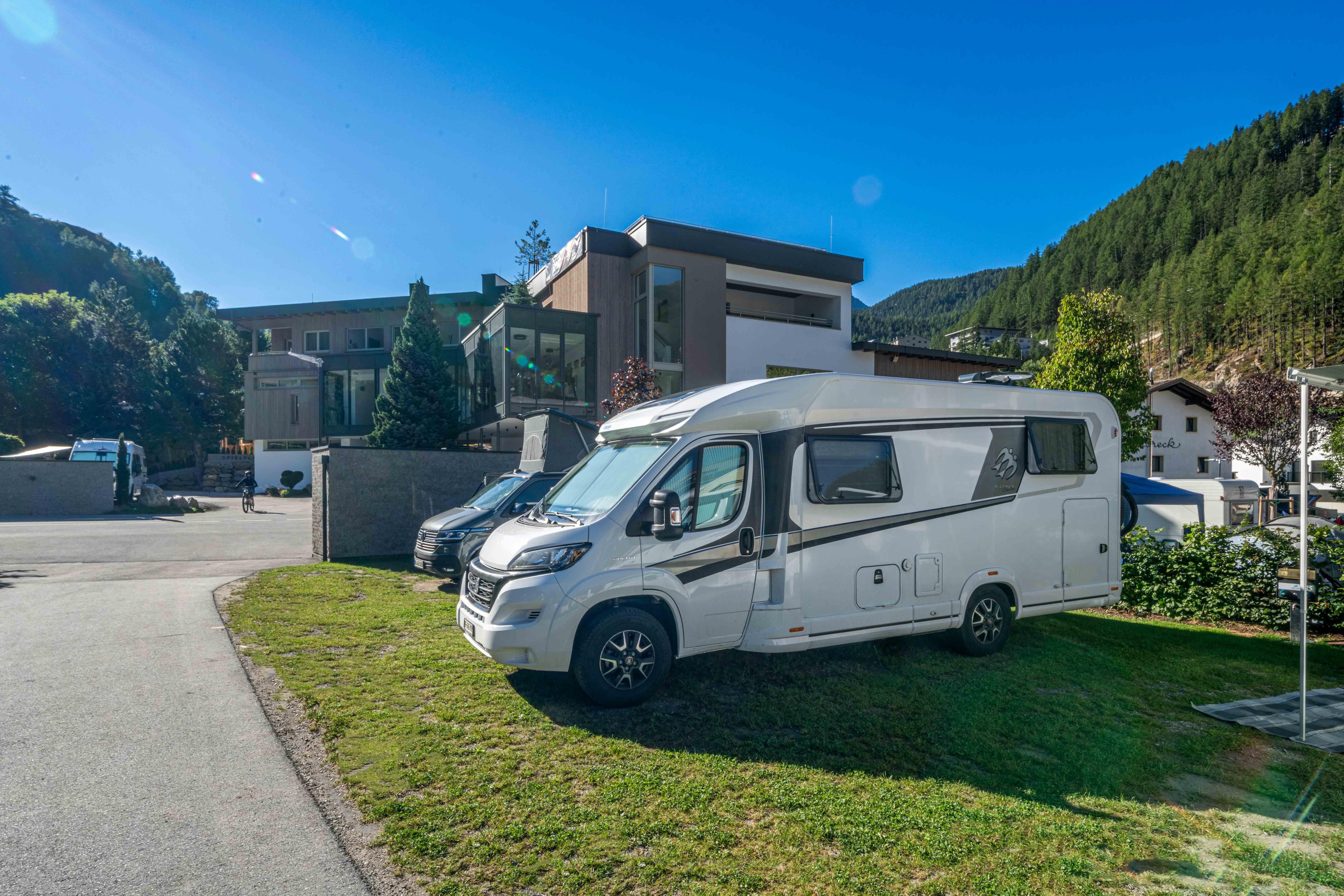 A motorhome is standing on a meadow in front of a modern building. In the background, mountains and a blue sky can be seen.