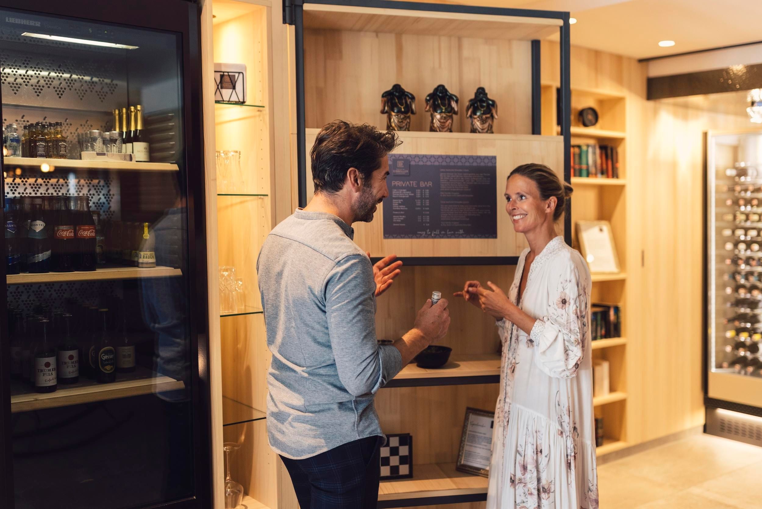 Two people are smiling while having a conversation in front of a shelf with various items. The surroundings have a warm, inviting atmosphere.