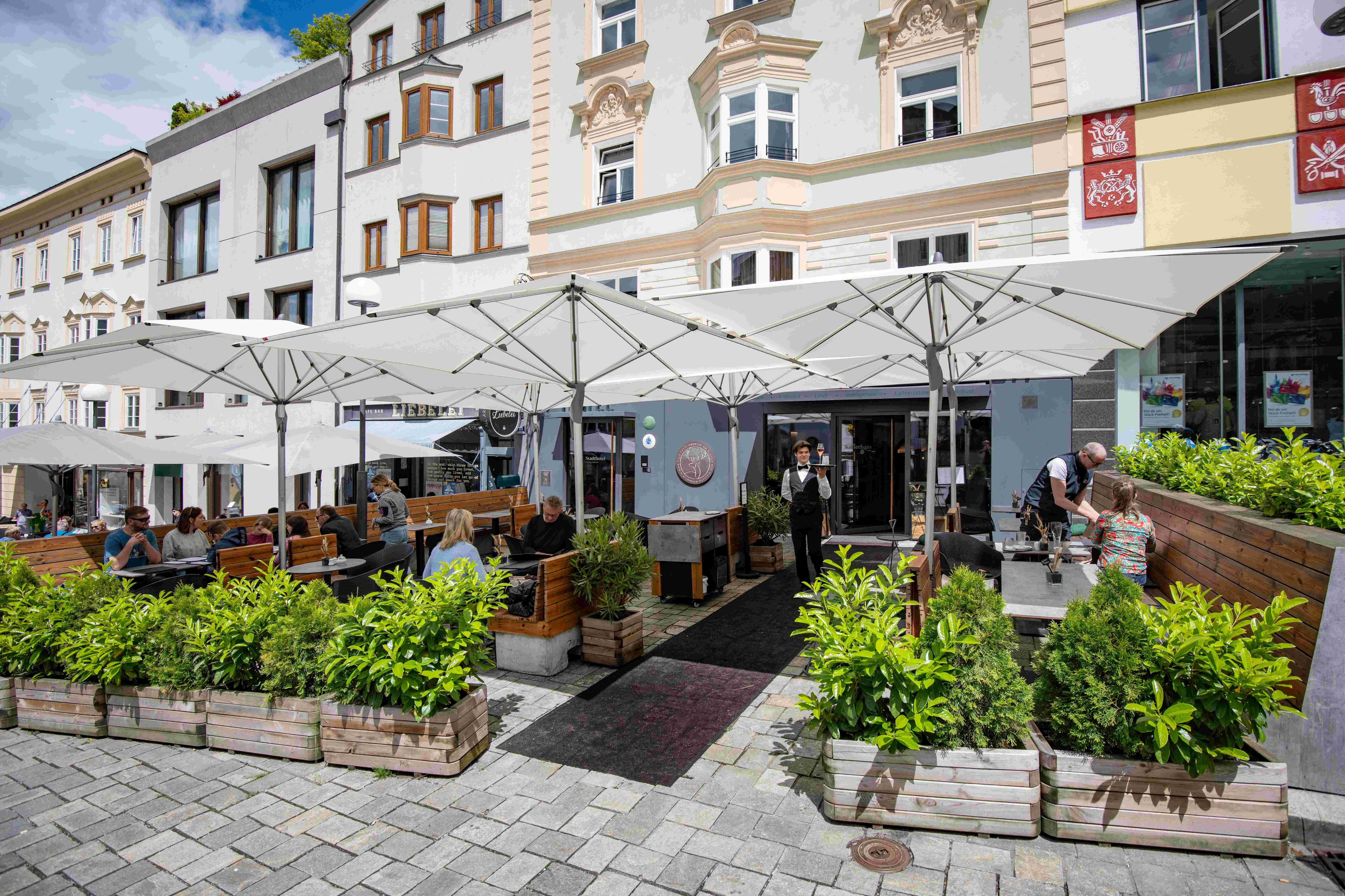 A cozy outdoor terrace of a café with sun umbrellas and plants. People are sitting relaxed and enjoying their time outside.