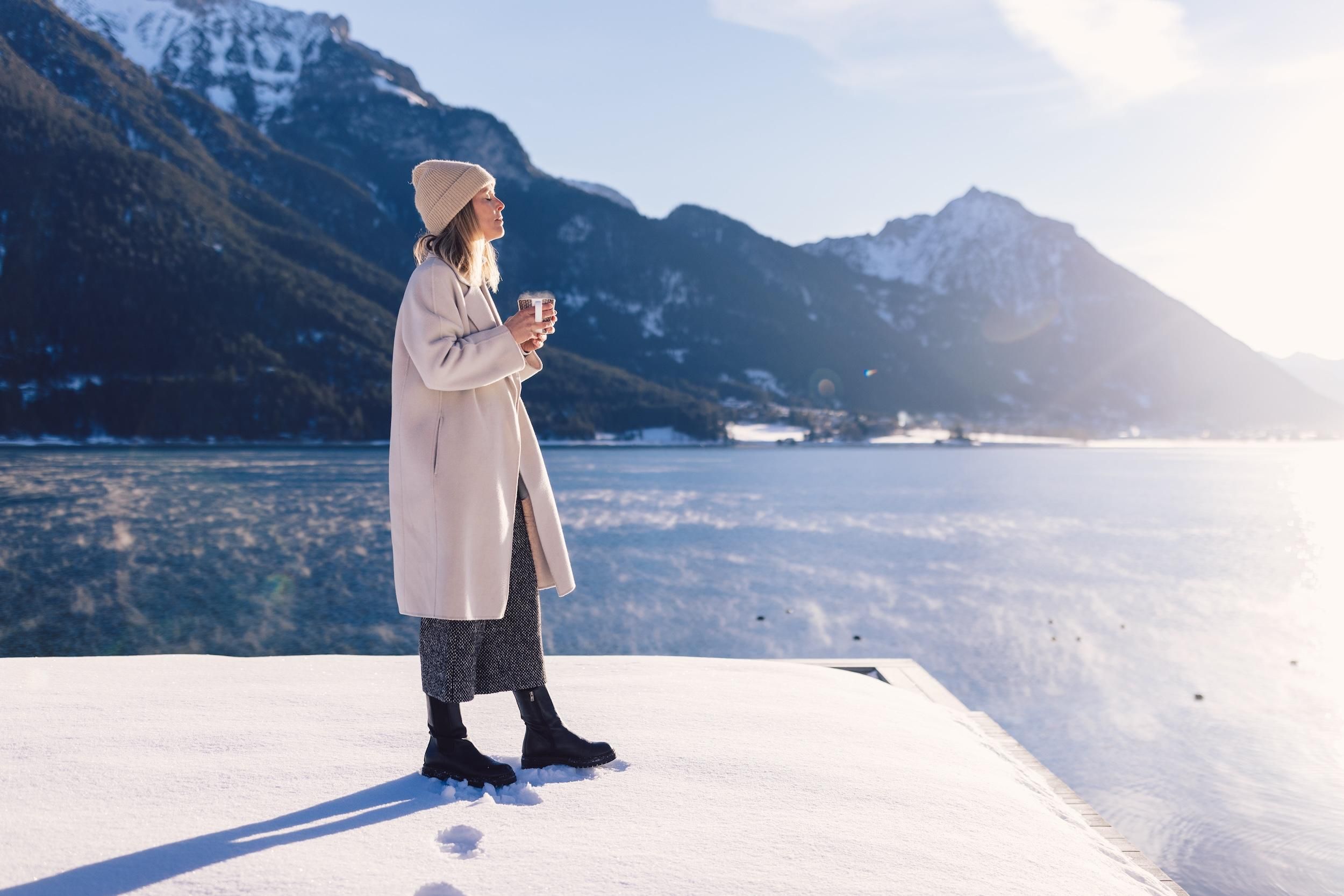 A woman stands on a snow-covered area, surrounded by mountains and a frozen lake. She holds a cup in her hand and enjoys the wintry landscape.