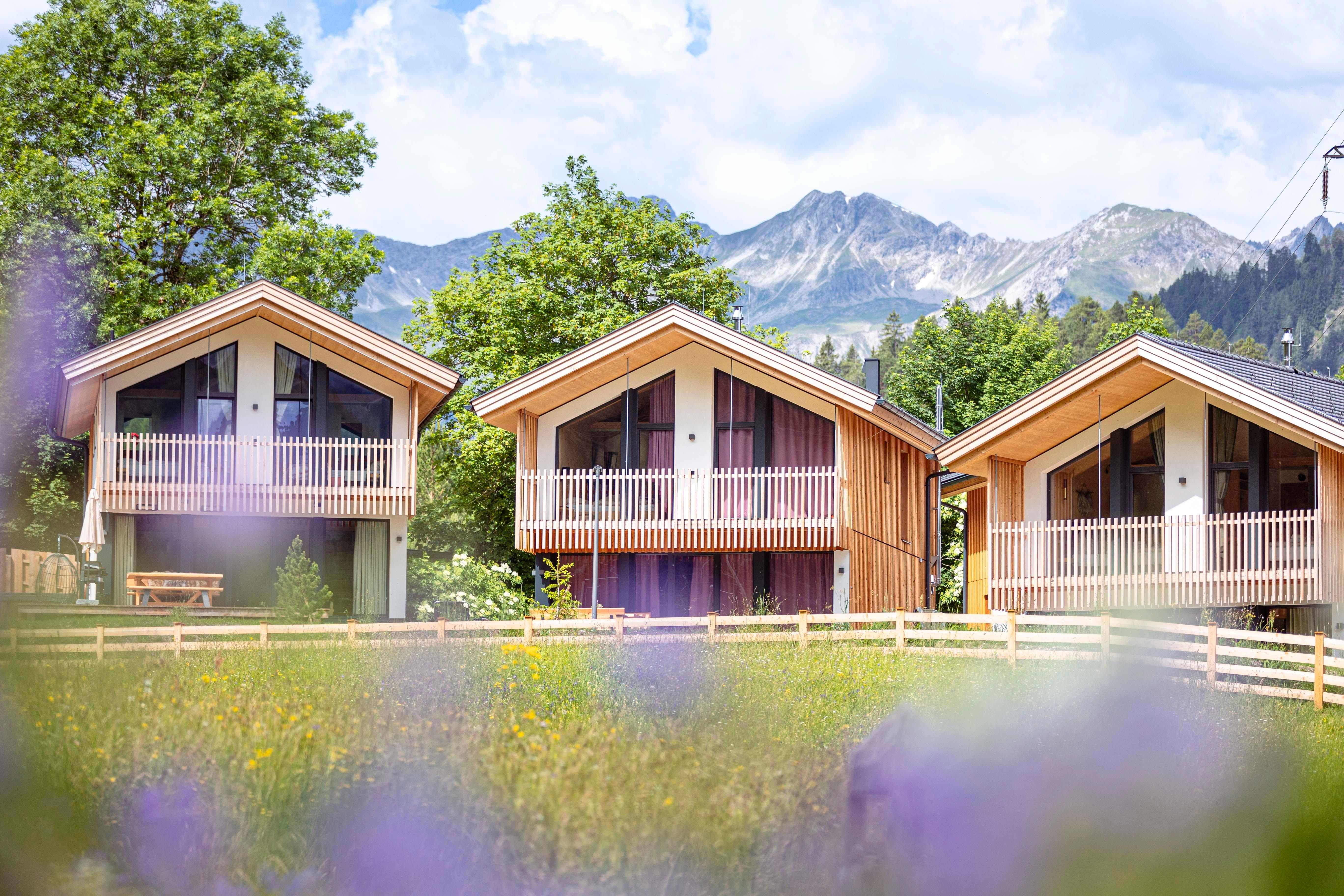 Three modern wooden cabins in nature, surrounded by green trees and mountains in the background. A beautiful, clear sky complements the idyllic landscape.