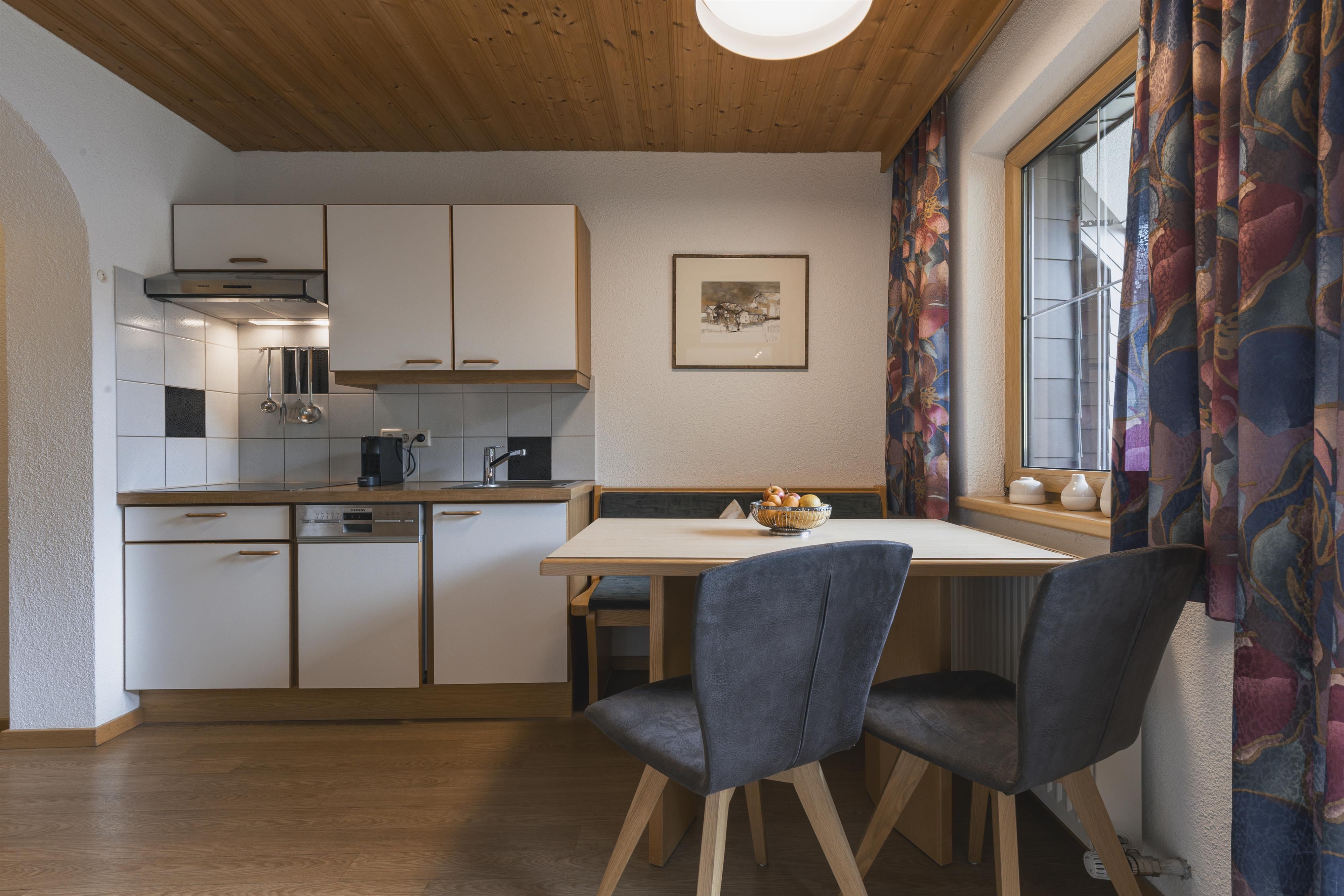 A modern kitchen with light furniture and a dining table. Daylight streams into the room through the window.