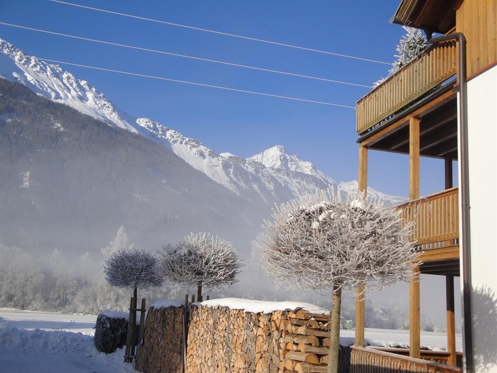A snowy mountain panorama with a clear blue sky. In the foreground, there is a stack of wood and frosty trees next to a wooden house.