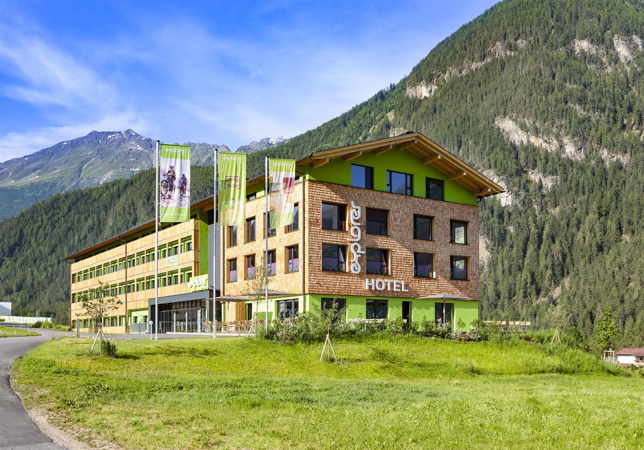 The Explorer Hotel Ötztal presents the exterior view in summer. In the background, the Ötztal Alps can be seen.