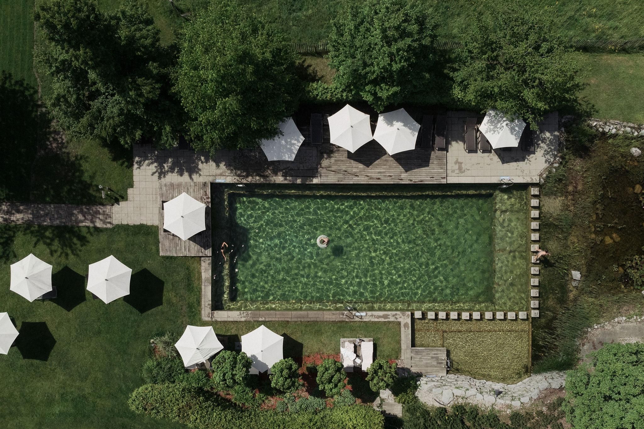 A beautiful pool surrounded by trees and lounge chairs. White sun umbrellas are on the terrace.