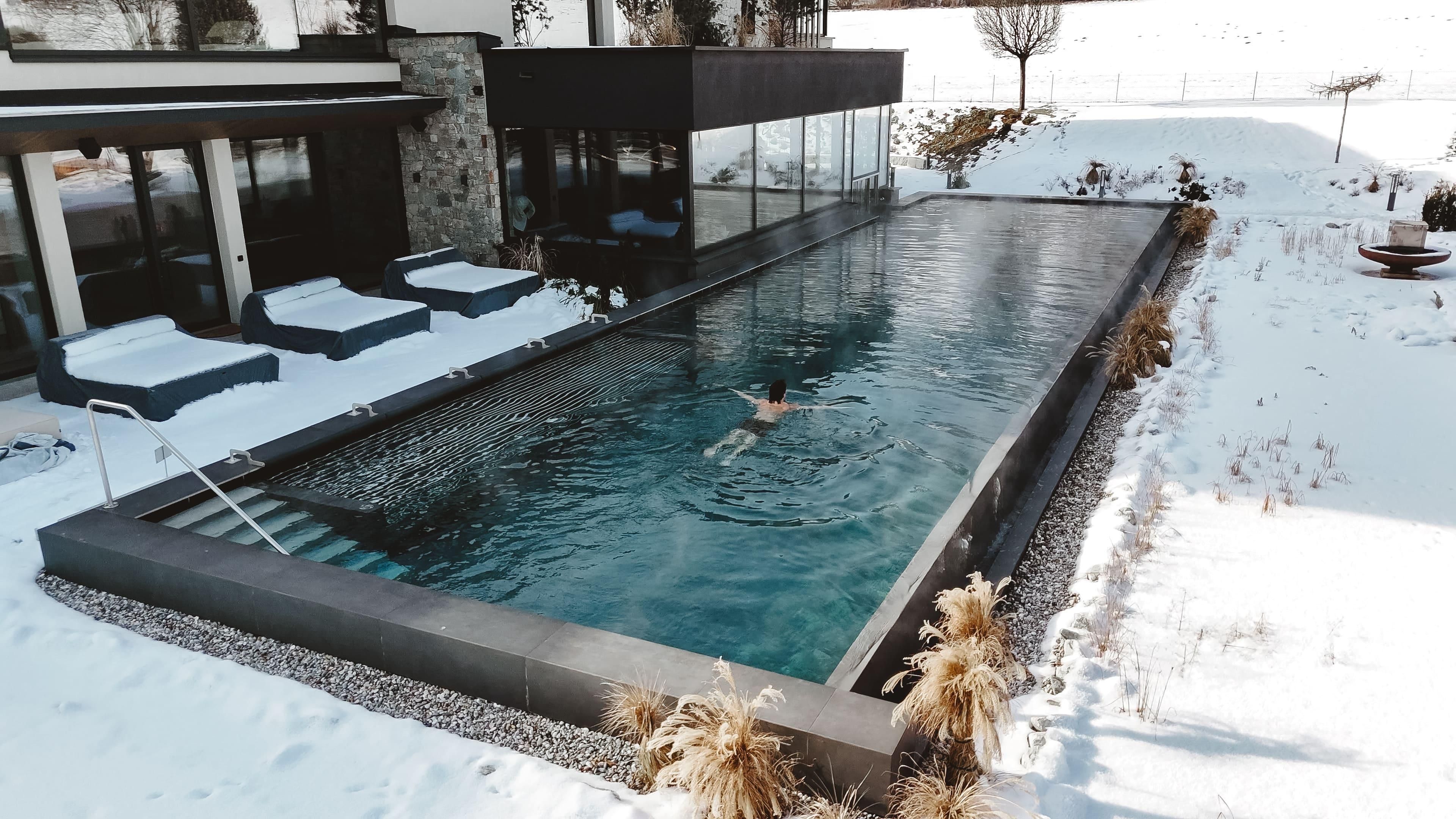 A modern pool, surrounded by snow, ideal for relaxation. One person swims in the clear water, while a calm winter landscape is visible in the background.