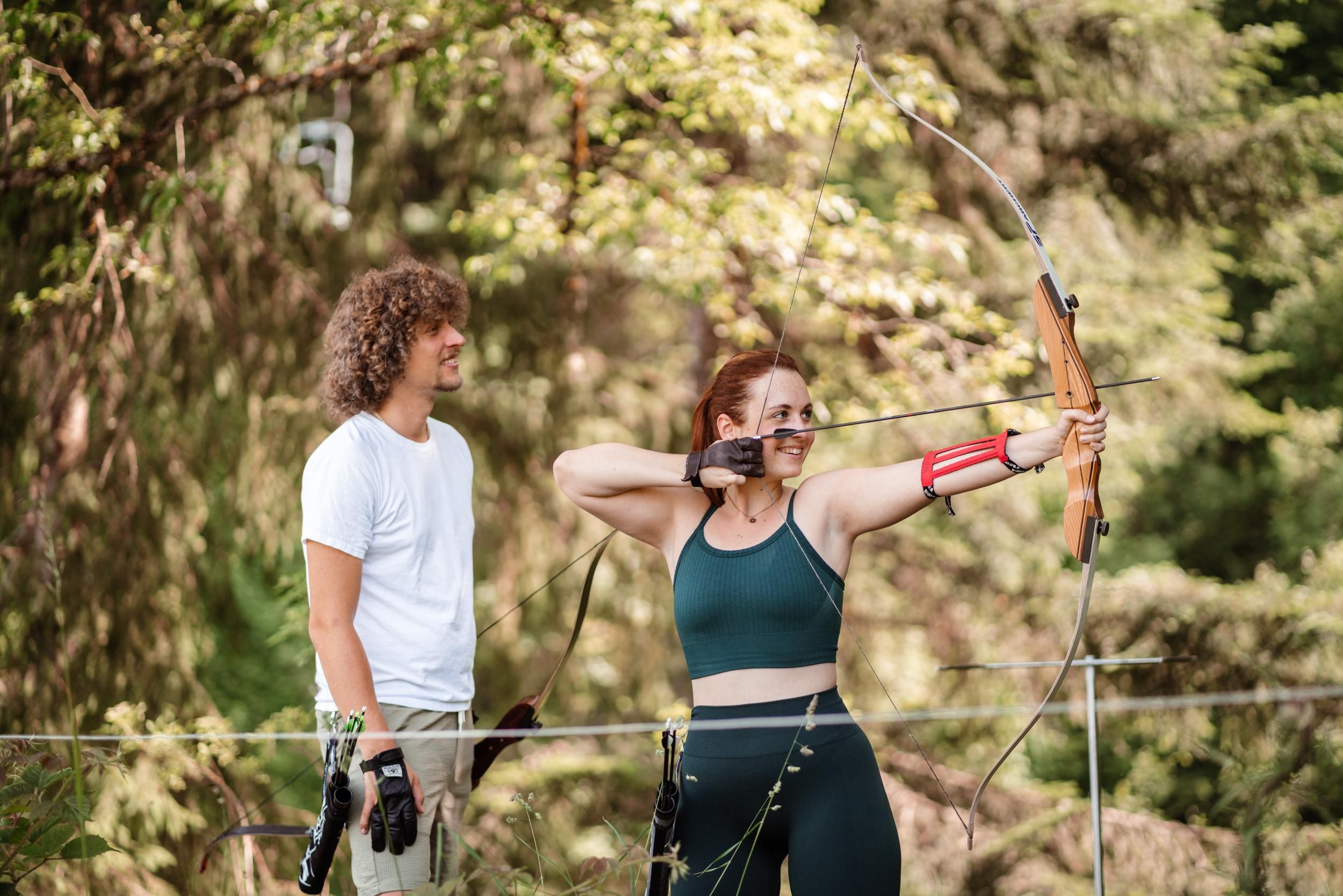A woman is shooting with a bow in nature, while a man watches her. Both are wearing sportswear and appear focused.