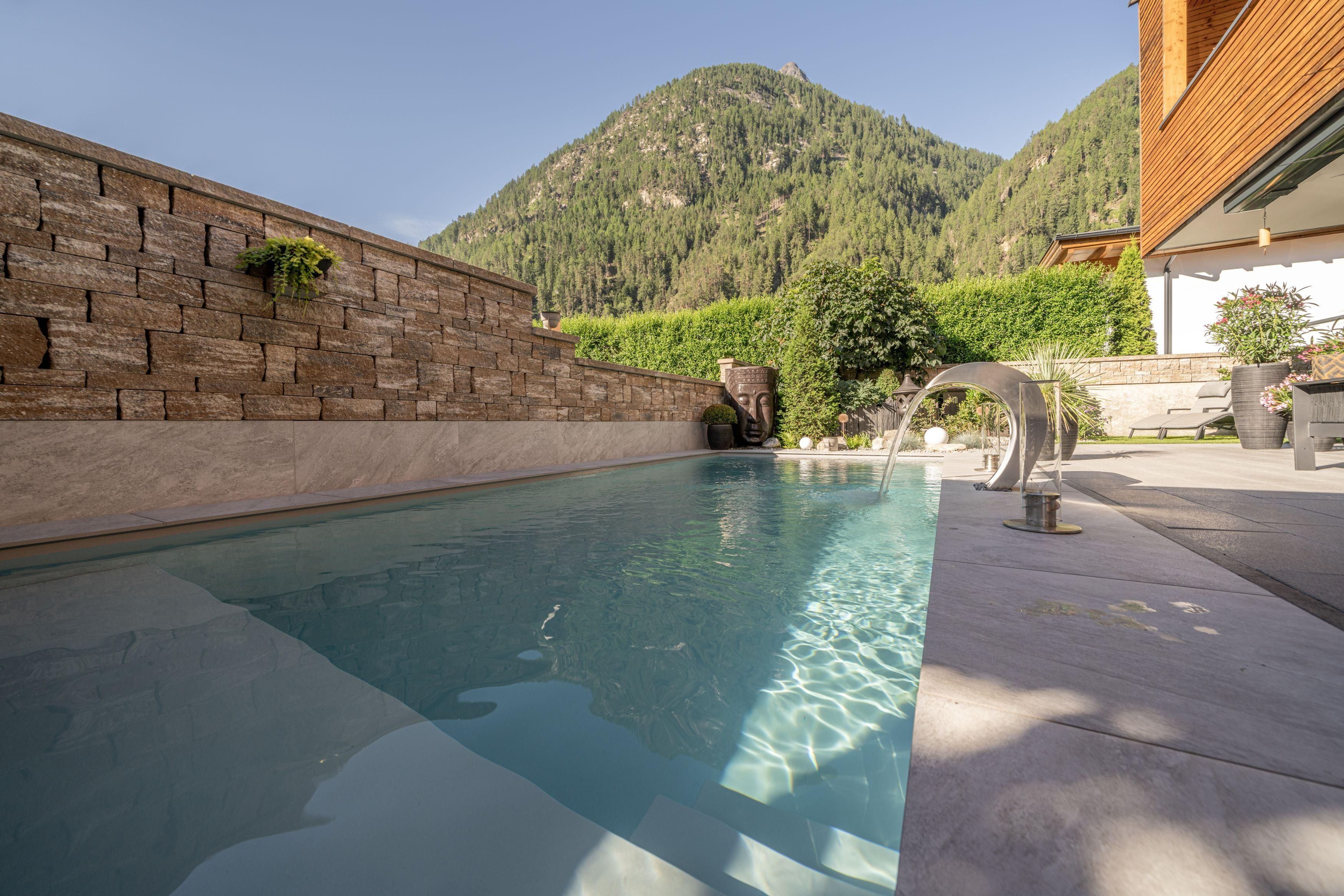 A modern pool surrounded by beautiful landscaping. In the background, green mountains are visible.