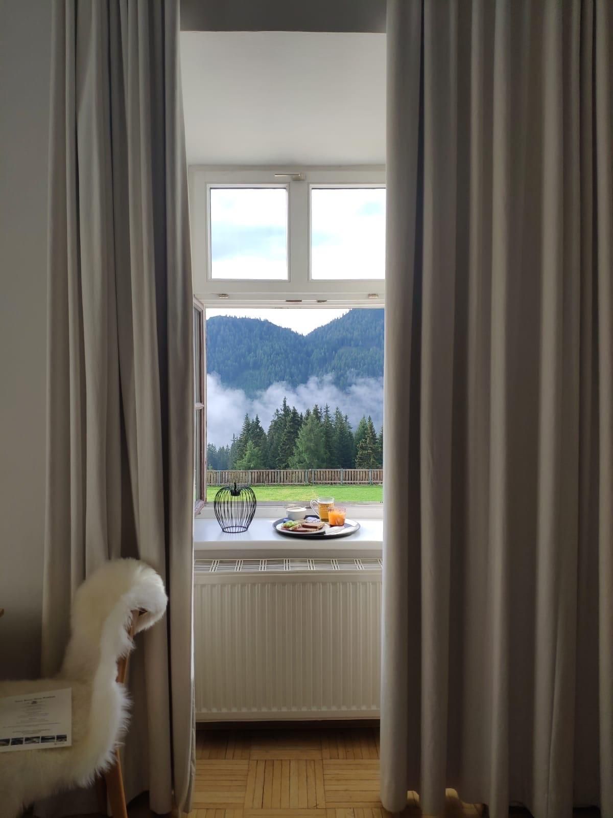 A cozy view from the window overlooking green mountains and clouds. On a table, fruits and a vase are visible.